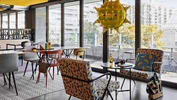 Espaço de estar interno moderno com mesas, cadeiras, janelas amplas e um balão decorativo em formato de sol.