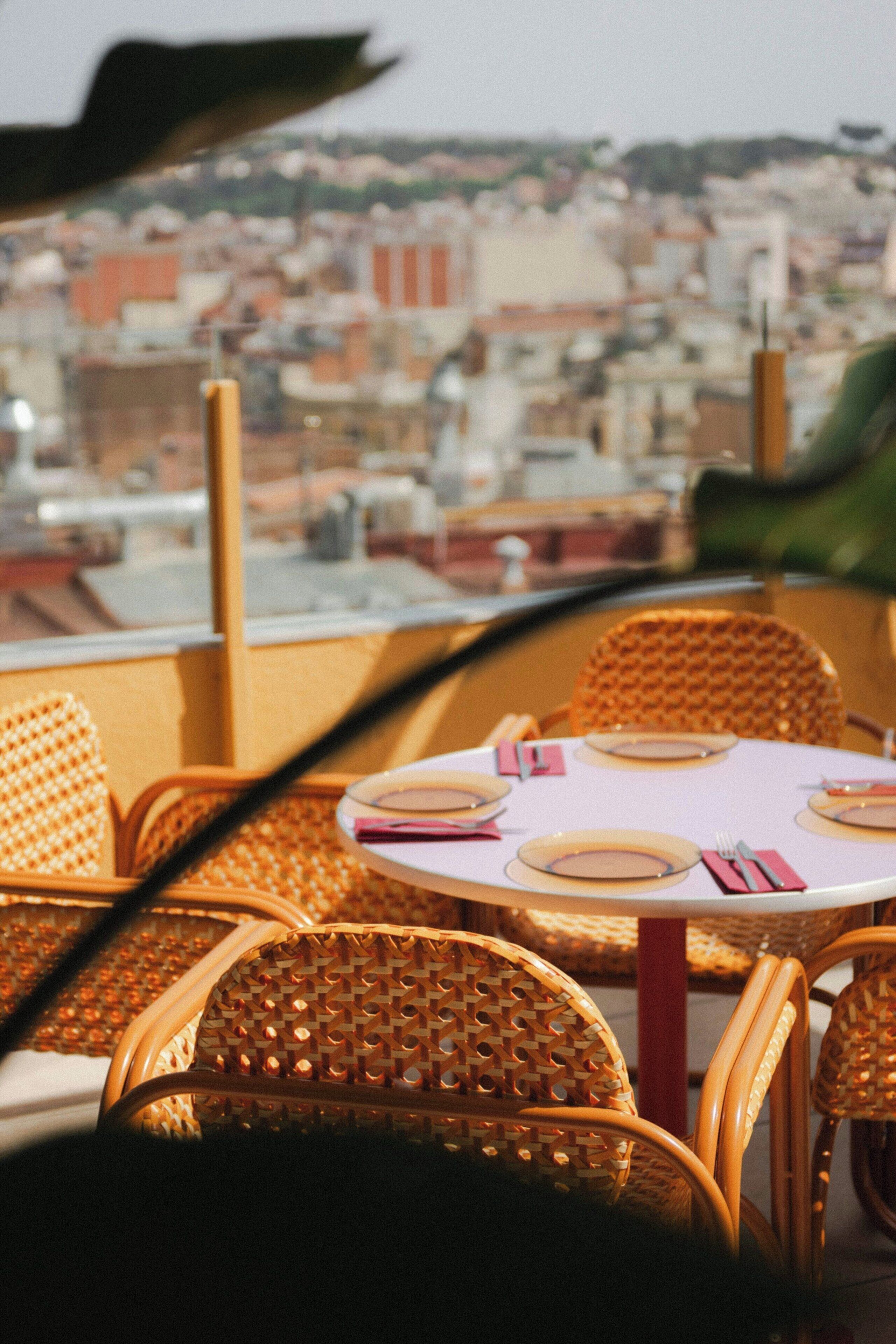 Una mesa puesta con platos, cubiertos y servilletas, con sillas de mimbre y vista a un paisaje urbano al fondo.