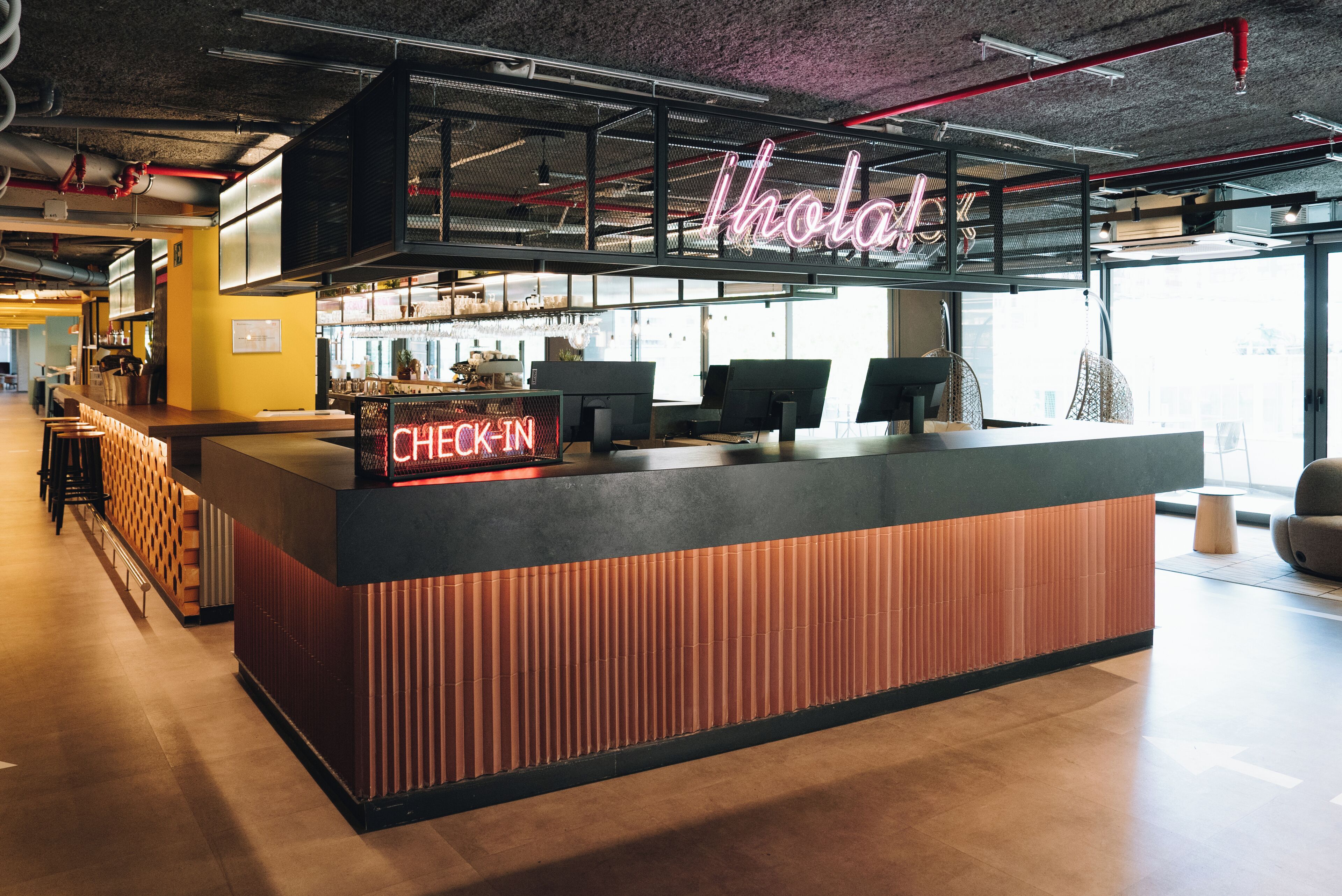 A modern check-in counter with a neon sign, a seating area, and a view of the city through large windows.