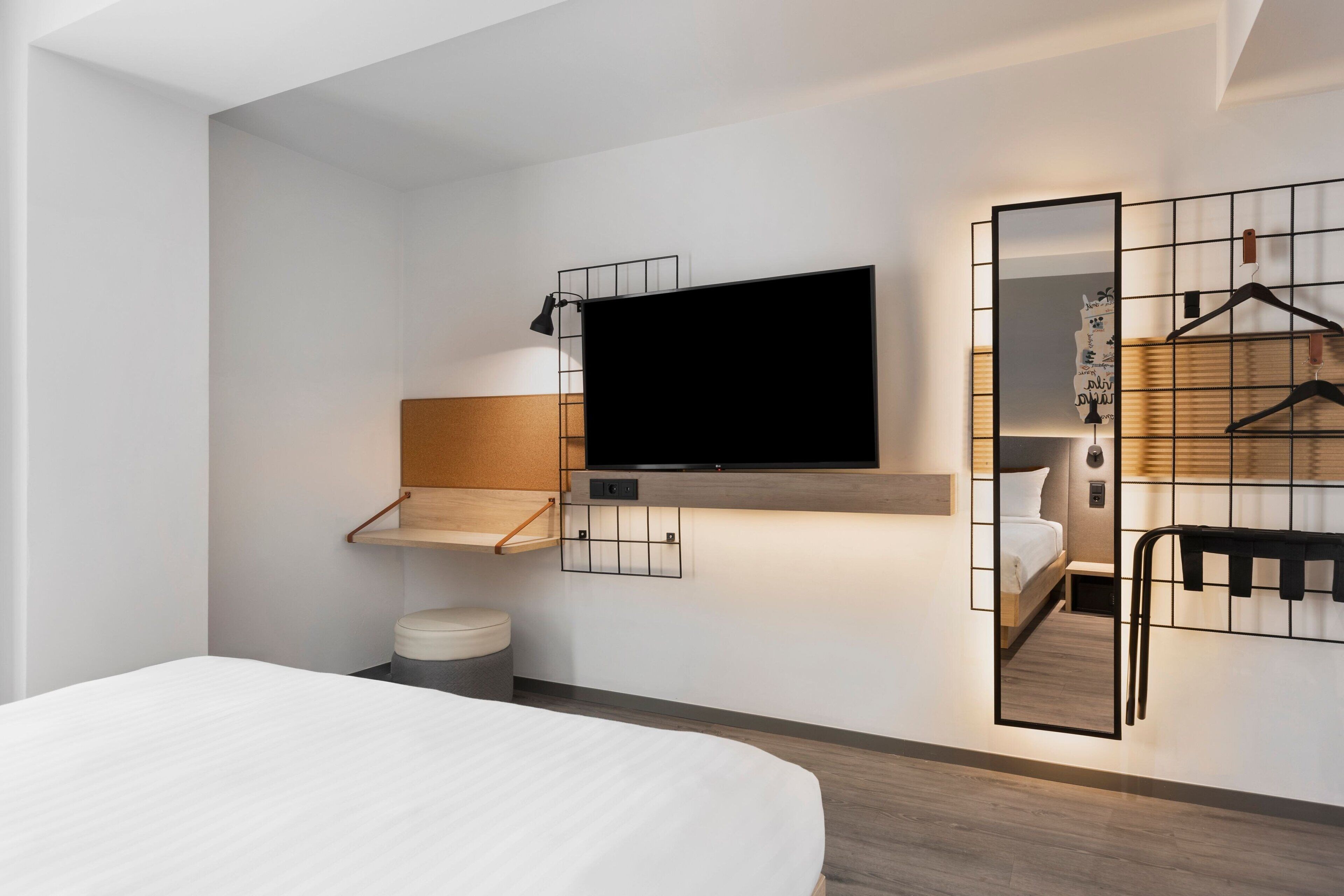 A modern hotel room with a large bed, a mounted TV, a wooden shelf, a mirror, and a hanging coat rack.