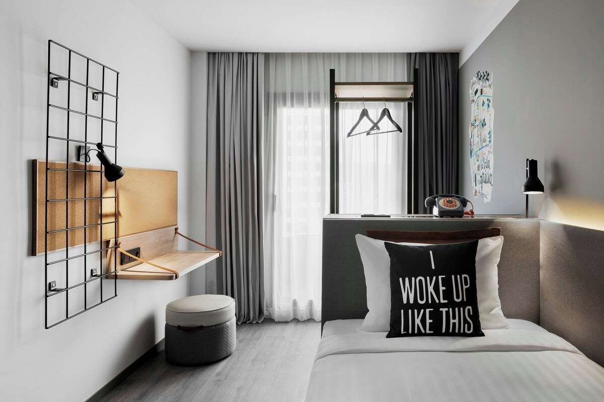 Une chambre d’hôtel moderne comprenant un lit, une table de chevet, une lampe suspendue, une fenêtre avec des rideaux et une étagère fixée au mur.