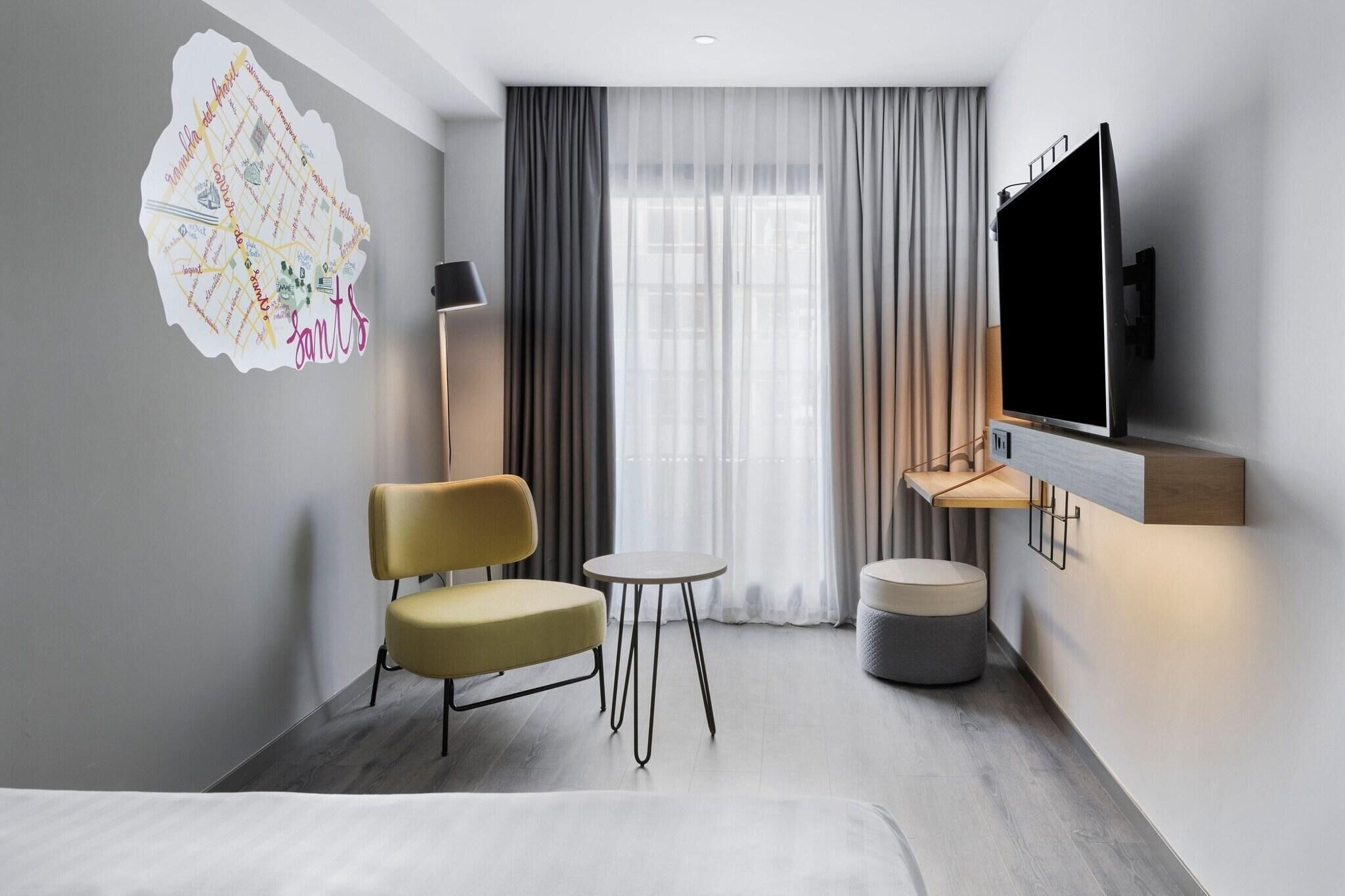 A modern hotel room with a bed, a yellow chair, a small round table, a TV mounted on the wall, and a large window with sheer curtains.