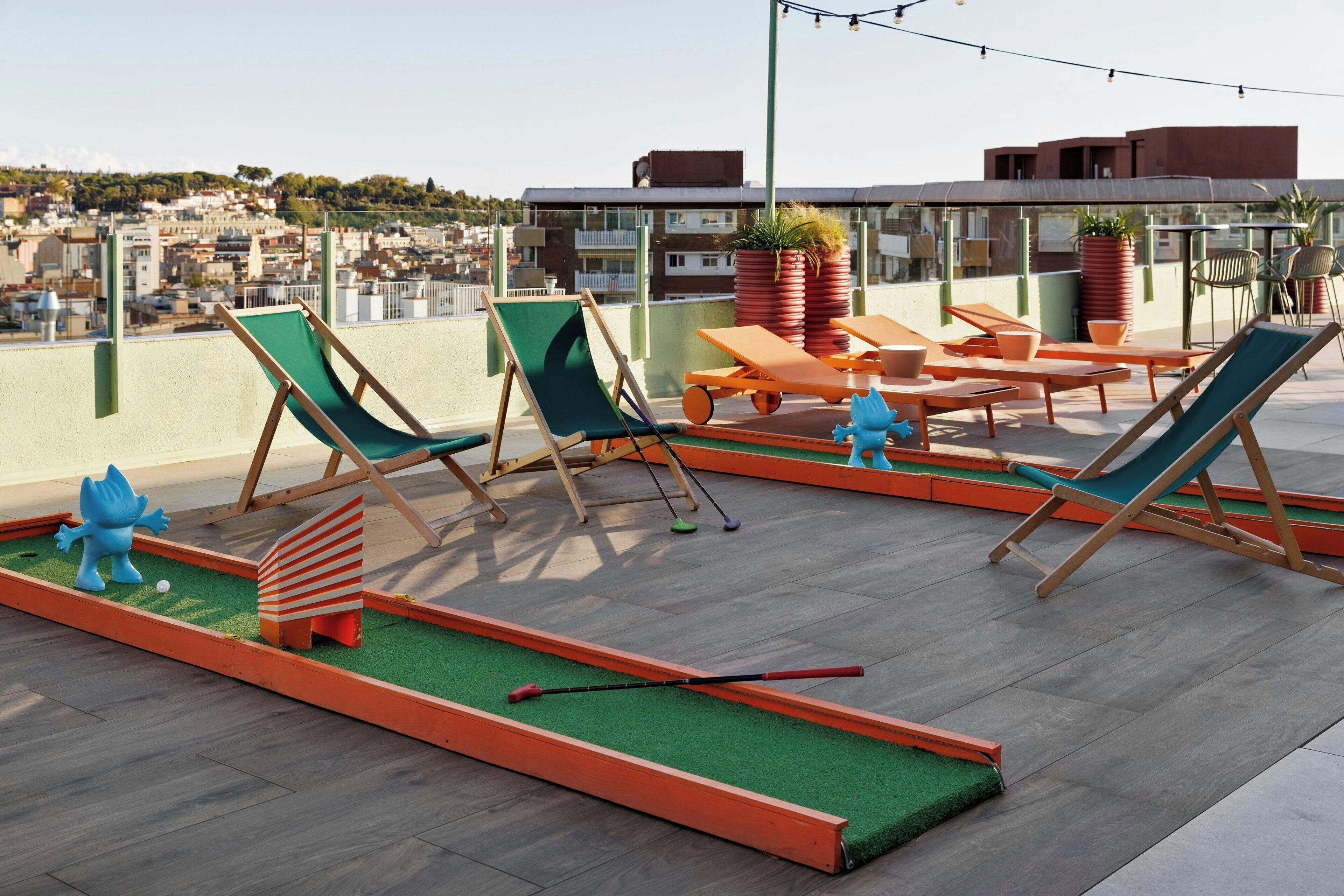 Un salón en la azotea con sillones verdes y naranjas, un pequeño campo de práctica de golf y vistas del paisaje urbano.