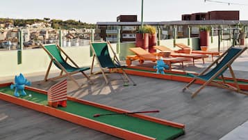 Un salón en la azotea con sillones verdes y naranjas, un pequeño campo de práctica de golf y vistas del paisaje urbano.