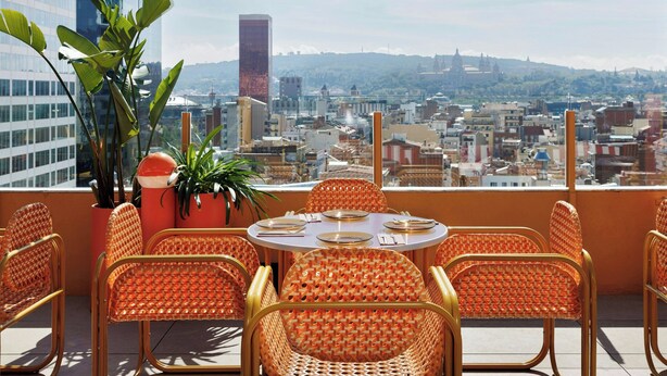 Una terraza en la azotea con sillas de mimbre y una mesa lista para comer, con vistas a un paisaje urbano que incluye edificios y una colina al fondo.