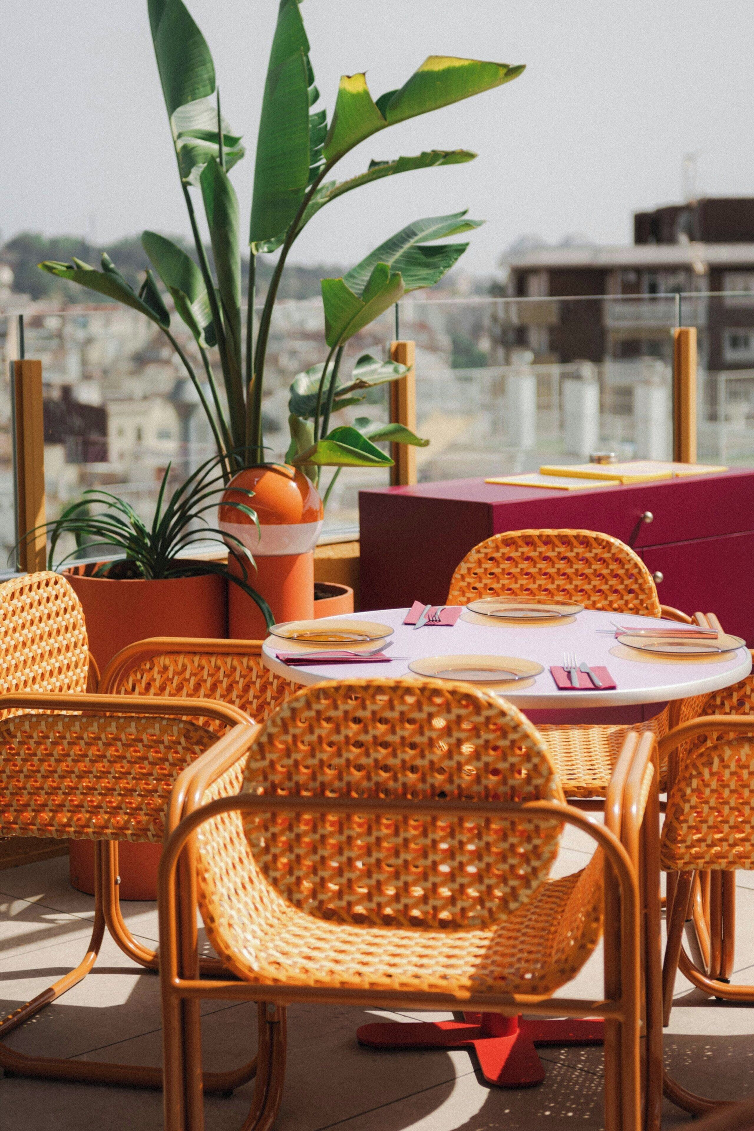 A dining area with wicker chairs and a table set for two, surrounded by potted plants.