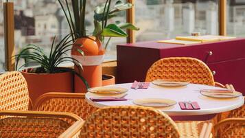 A dining area with wicker chairs and a table set for two, surrounded by potted plants.