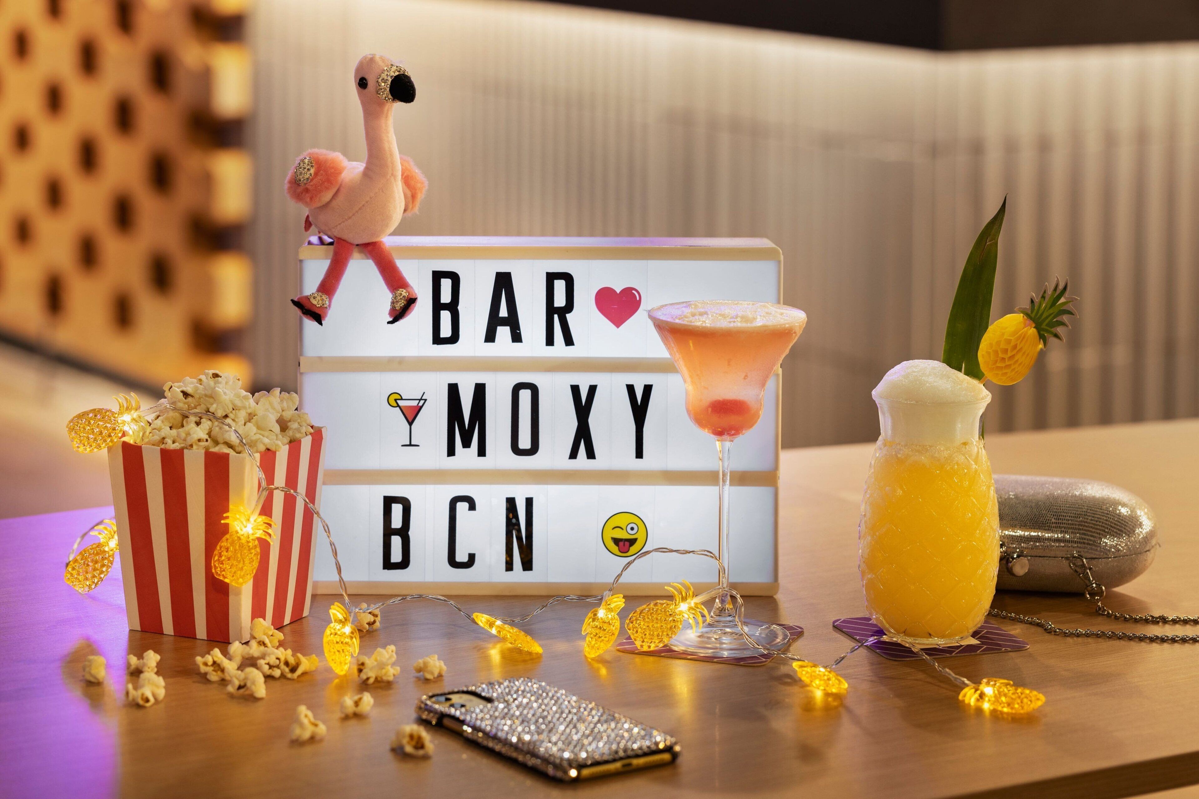 A bar setup with a sign reading "BAR MOXY BCN," a plush flamingo, a martini glass, a pineapple drink, a striped popcorn bucket, and a smartphone on a wooden table.