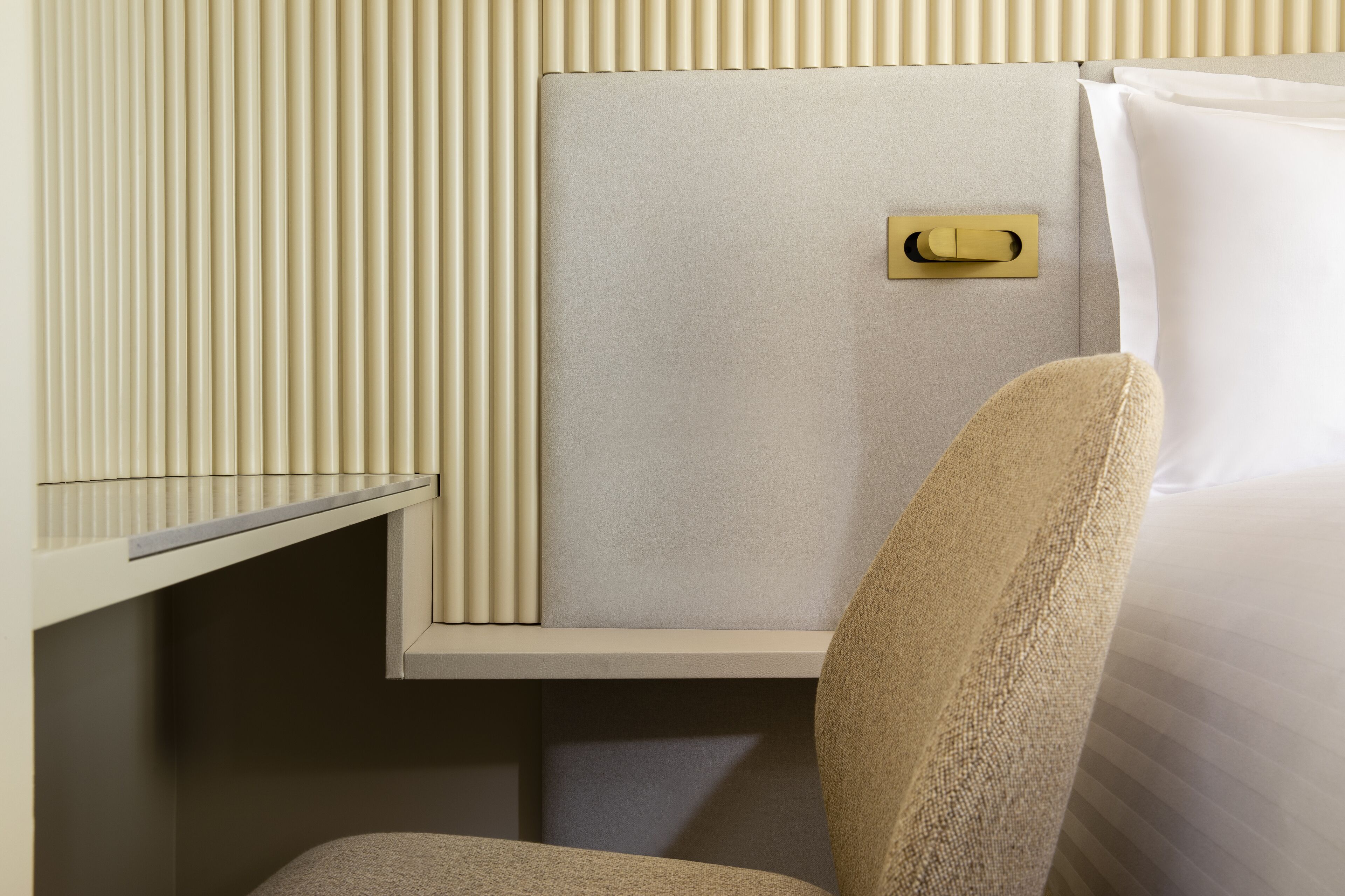 A hotel room with a bed, a bedside table, a chair, a wall panel, and a light switch.