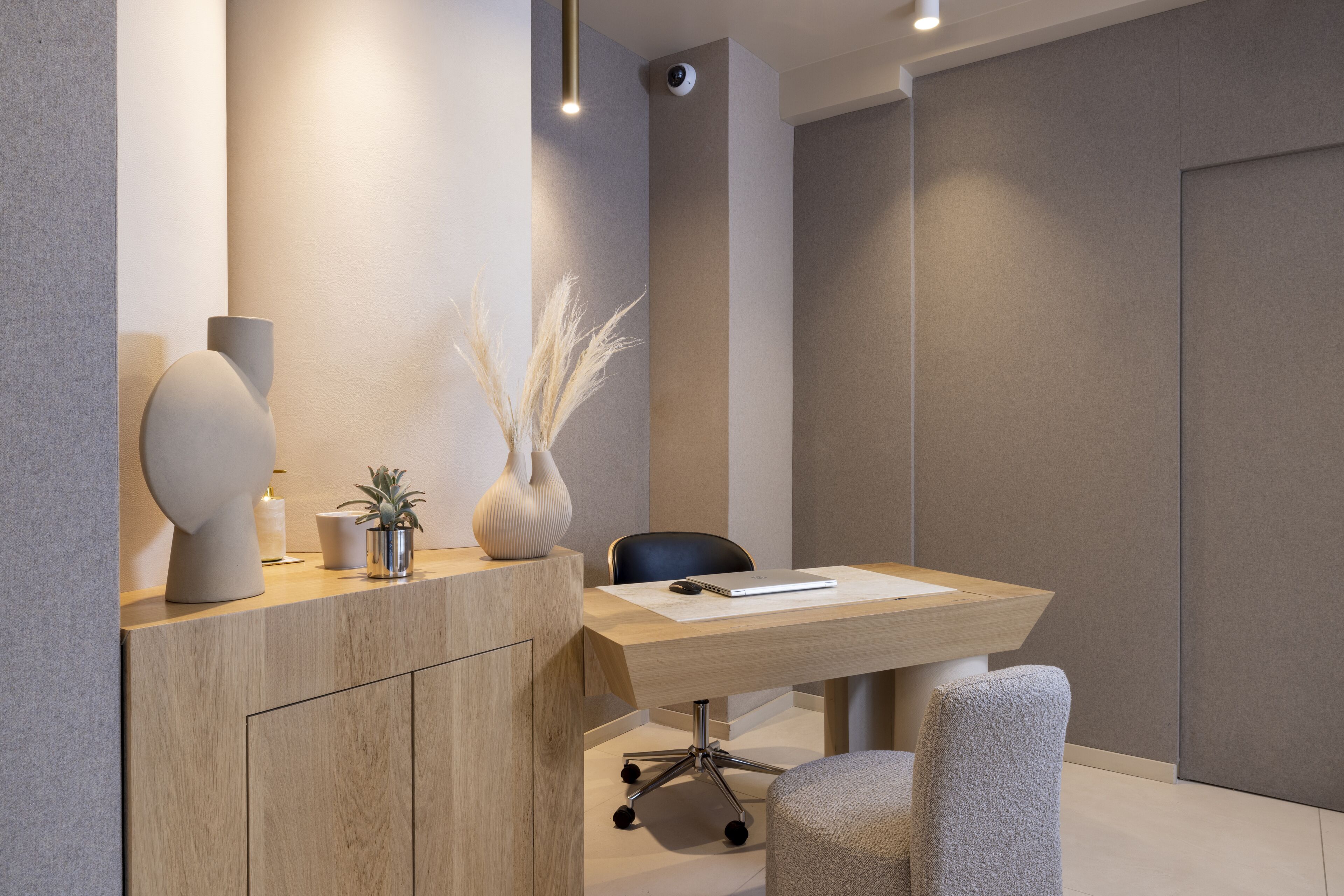 Un espace de bureau moderne avec un bureau en bois, une chaise de bureau noire, un vase contenant des plantes séchées et une caméra de surveillance fixée au mur.