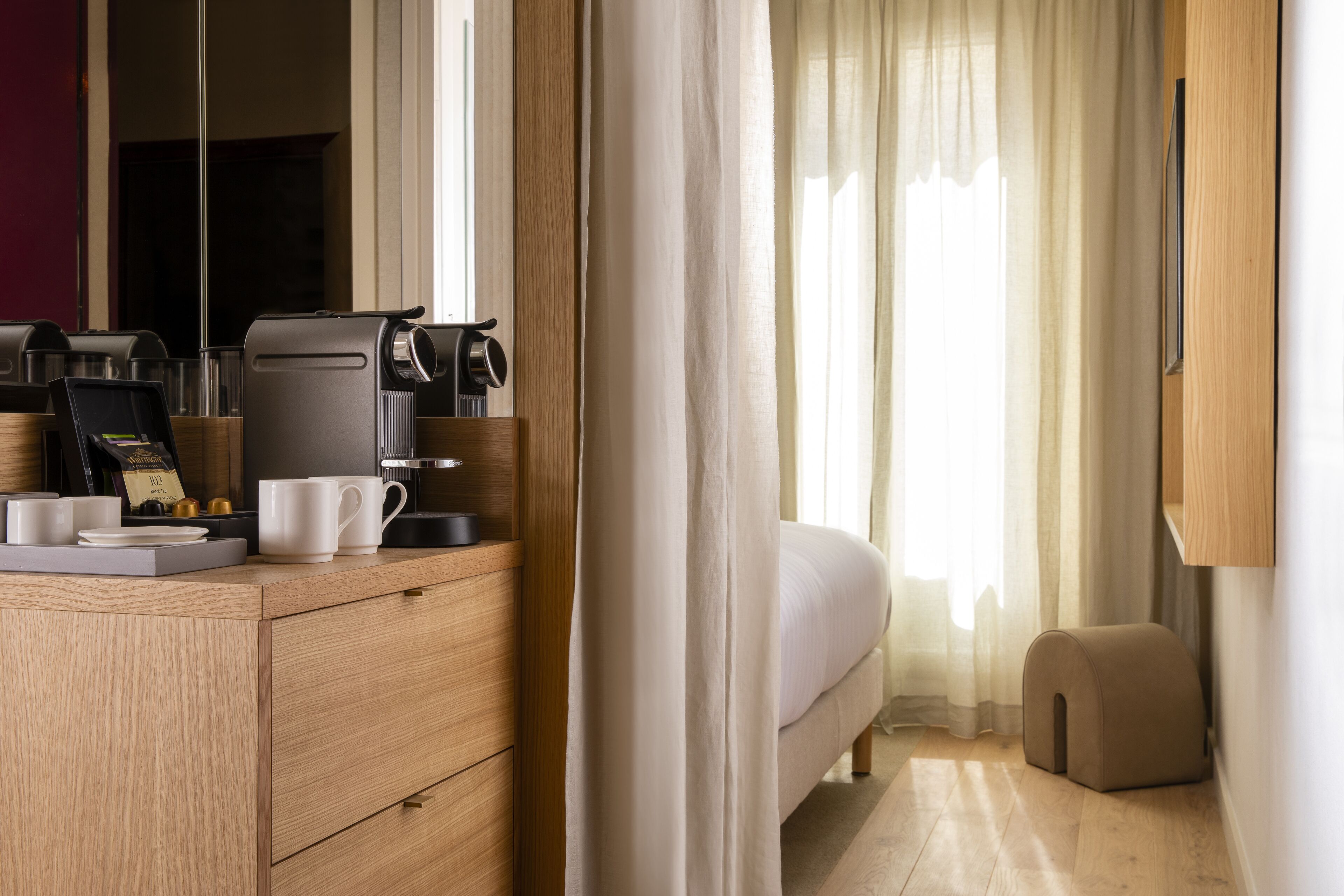 A hotel room with a wooden dresser, a bed, a bedside table with a coffee maker, and a small ottoman.