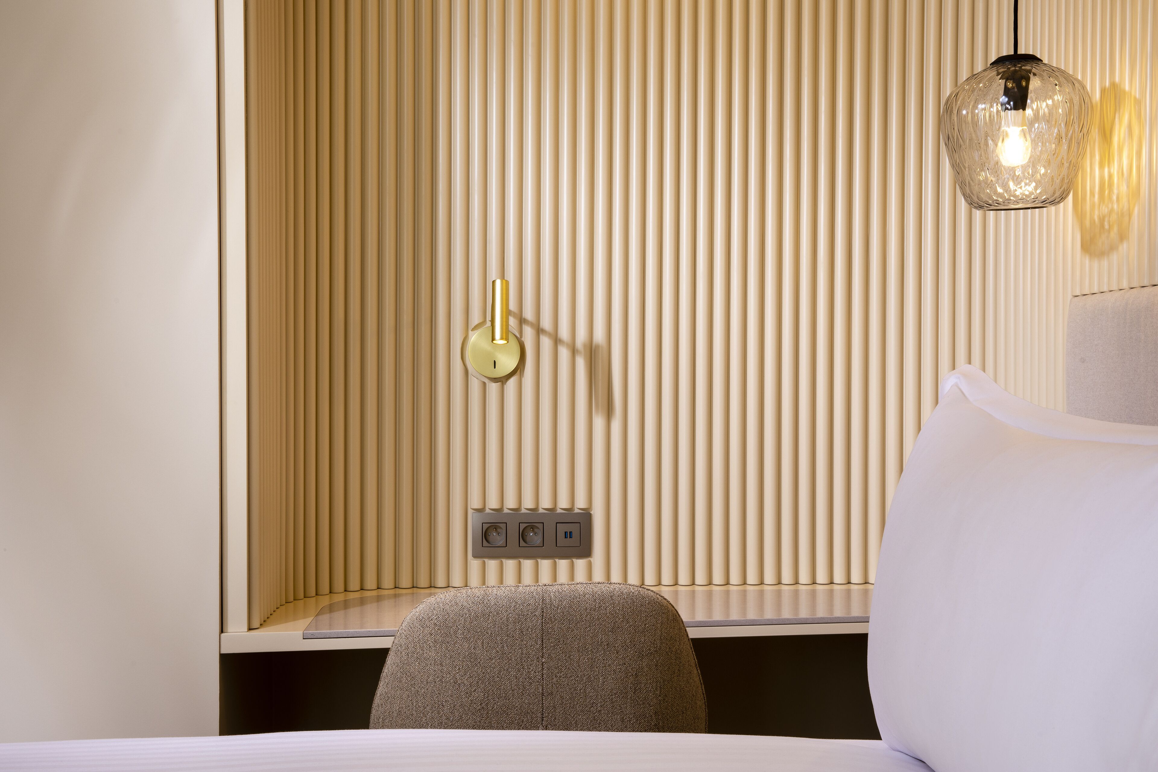 A hotel room with a bed, a chair, a wall-mounted light fixture, and a power outlet.