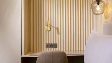 A hotel room with a bed, a chair, a wall-mounted light fixture, and a power outlet.