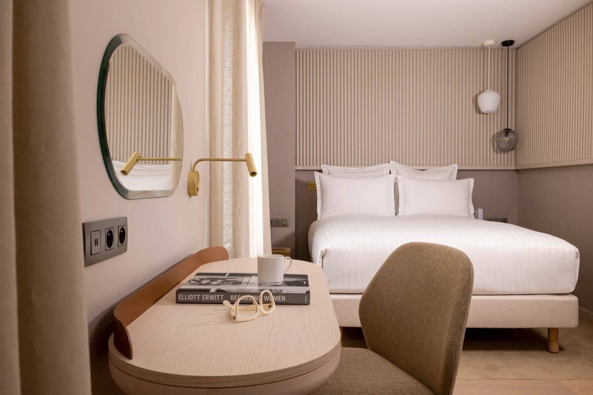Une chambre d’hôtel avec un lit, un bureau et une chaise, un miroir et une table de chevet avec un livre et des lunettes.