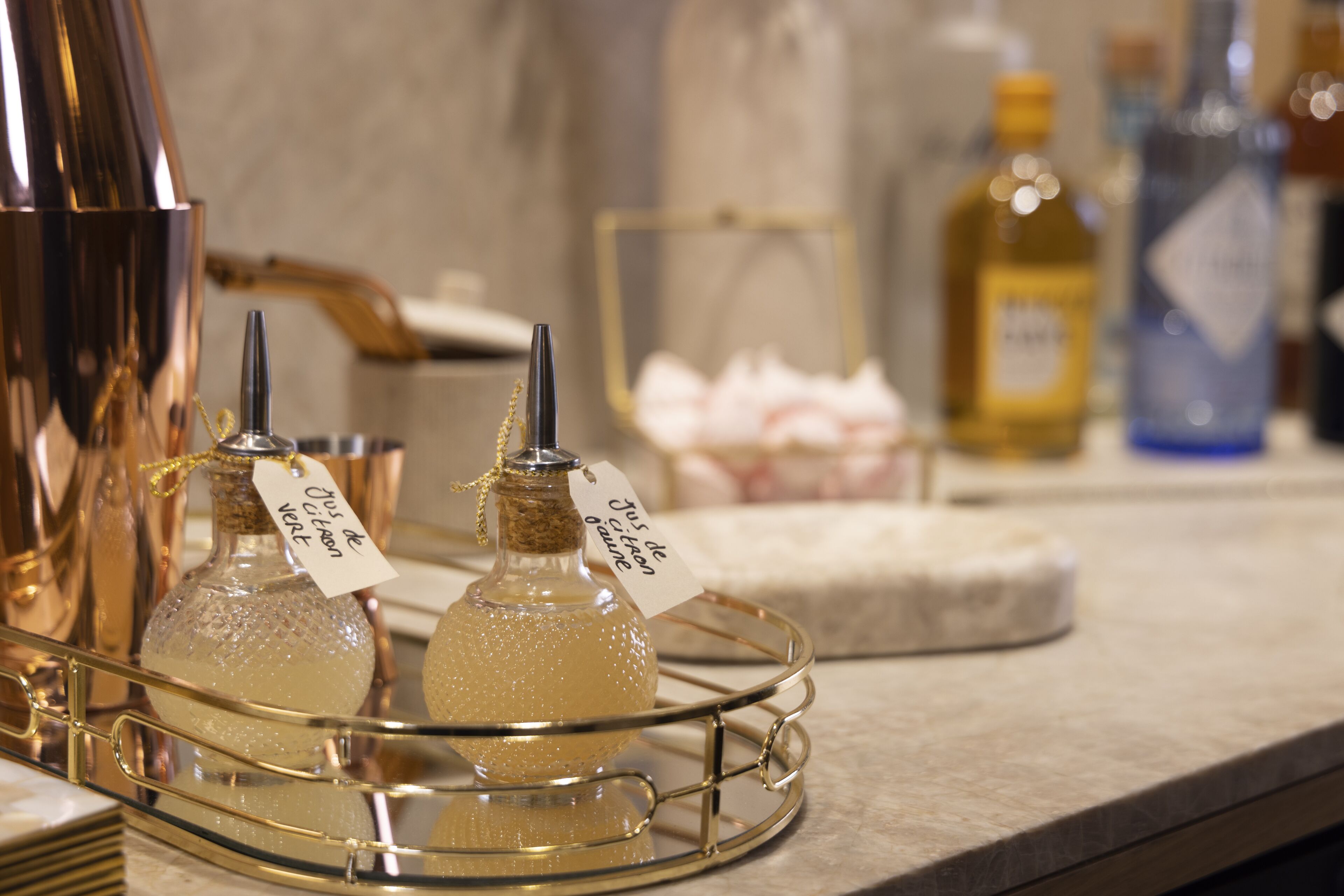 A bar area with a copper shaker, two bottles of a clear liquid with tags, a bottle of olive oil, and a bottle of blue liquid on a marble countertop.