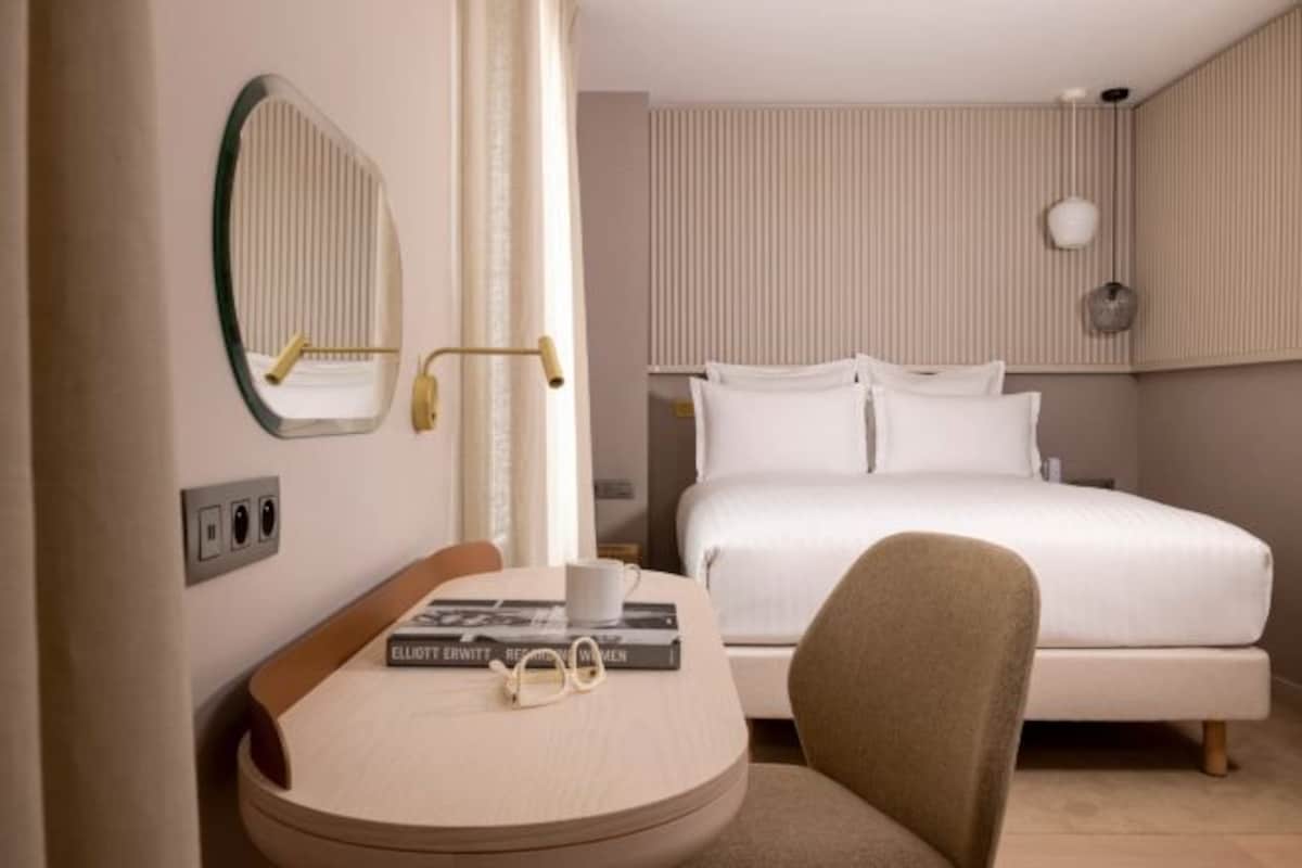 A hotel room with a bed, a desk with a chair, a mirror, and a bedside table with a book and glasses.