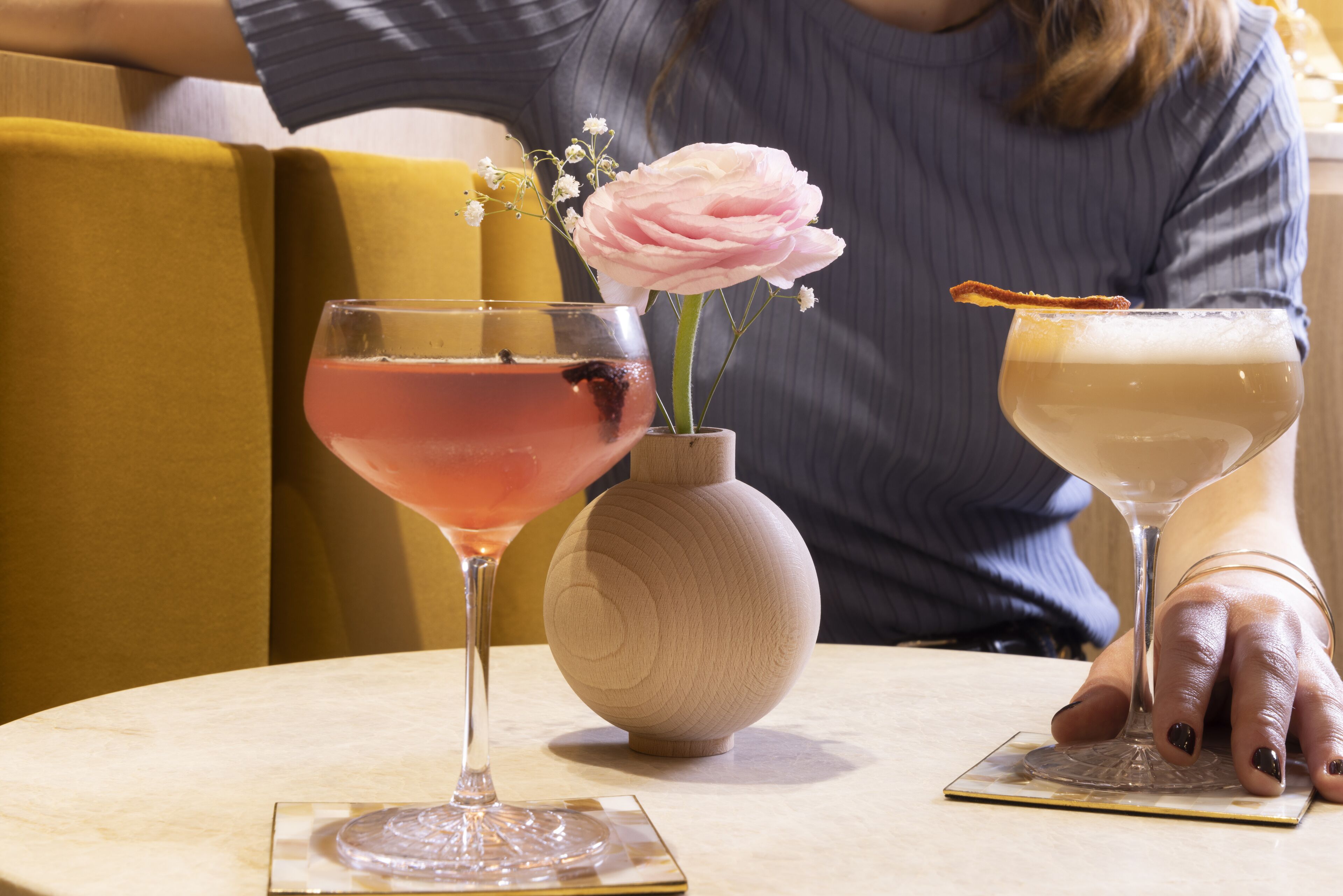 A table with two drinks, a vase with a flower, and a person sitting in the background.