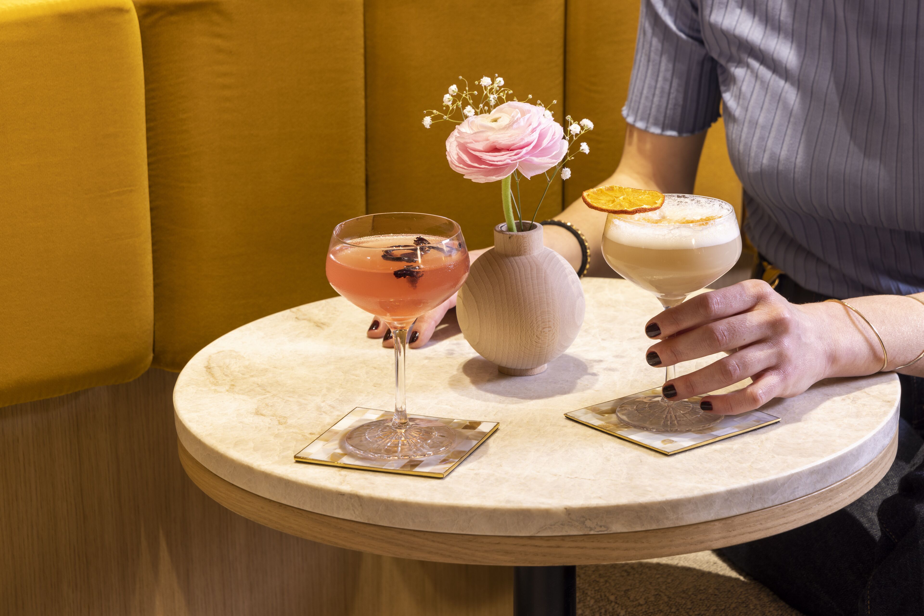 A round table with two drinks, a vase with flowers, and a coaster.