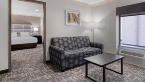 A hotel room with a grey patterned sofa, a small table, a bed with white bedding, a framed picture on the wall, a floor lamp, and a window with blinds. - Best Western Webster Hotel, Nasa (Webster)