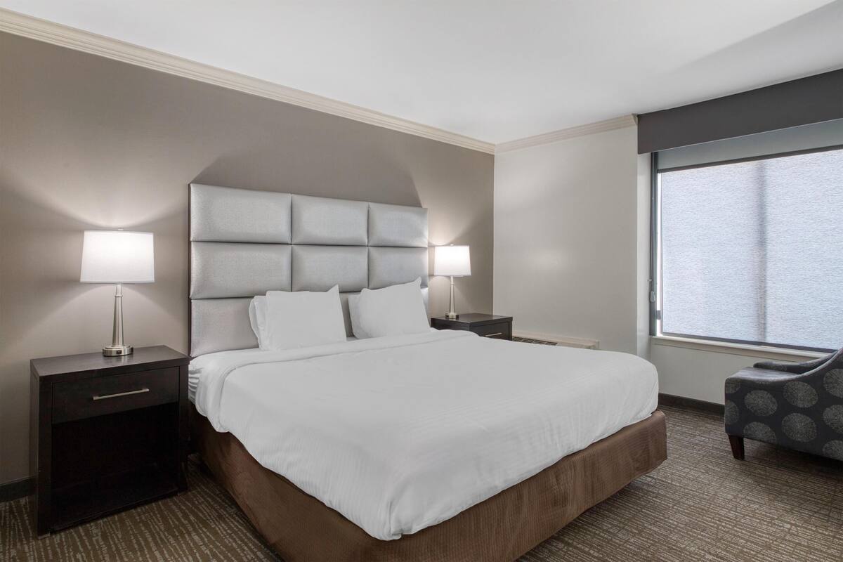 A hotel room with a large bed, two bedside tables with lamps, a chair, and a large window.