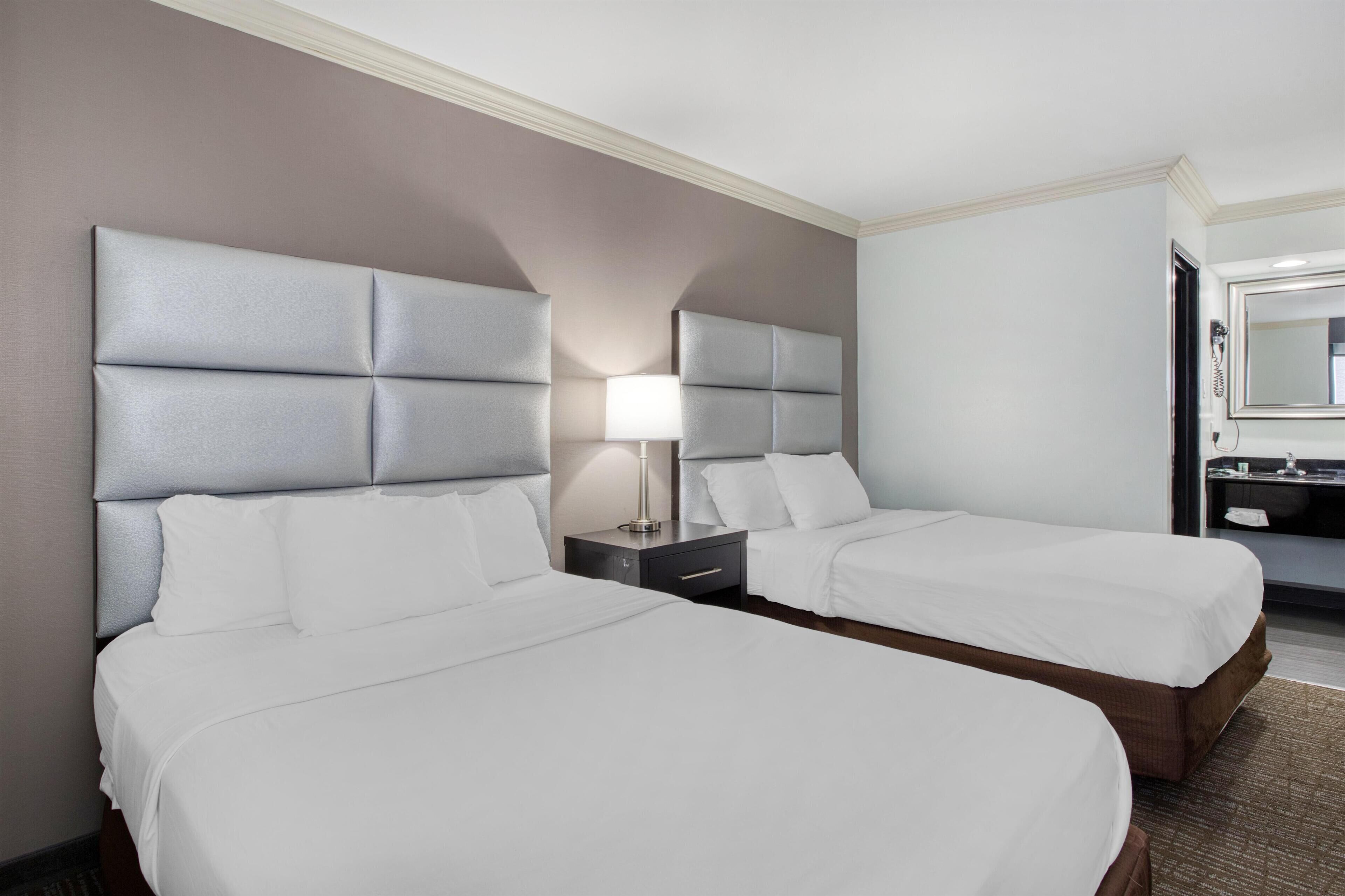 A hotel room with two beds, a nightstand with a lamp, a bathroom with a mirror, and a small table.