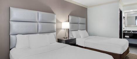 A hotel room with two beds, a nightstand with a lamp, a bathroom with a mirror, and a small table.