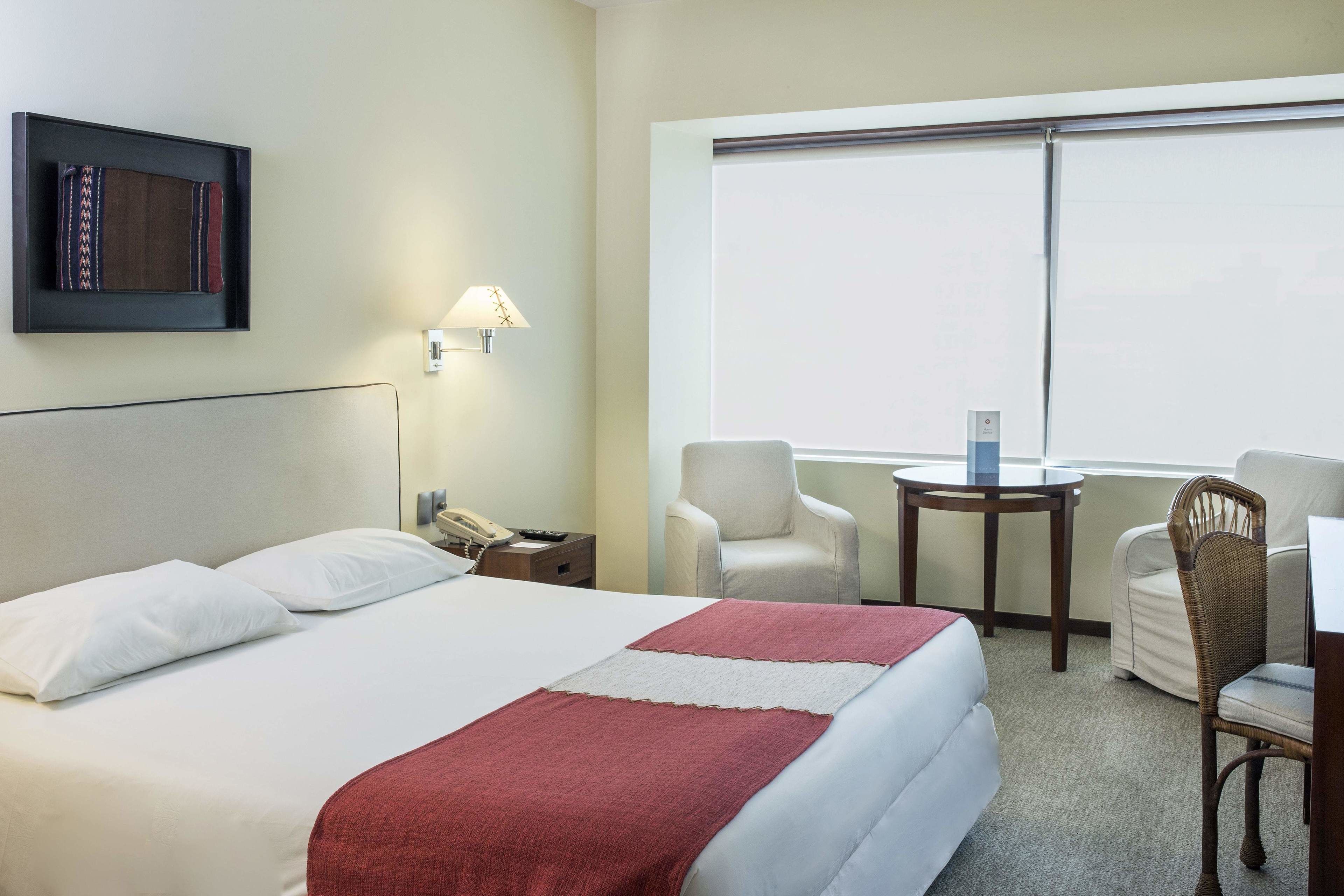 A hotel room with a bed, a desk, two chairs, a lamp, and a window.