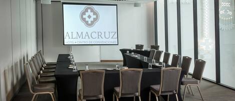 Un salón de conferencias con una larga mesa y sillas, una pantalla que muestra 'ALMACRUZ HOTEL CENTRO DE CONVENCIAS', y ventanas grandes con luces decorativas.