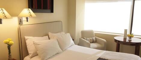 A hotel room with a bed, bedside lamps, a chair, a small table, and a window with curtains.