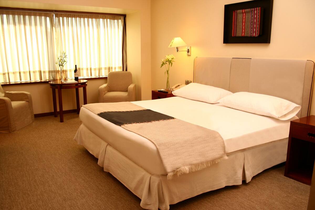 Une chambre d’hôtel avec un grand lit, une chaise, une petite table, une lampe et un tableau encadré au mur.