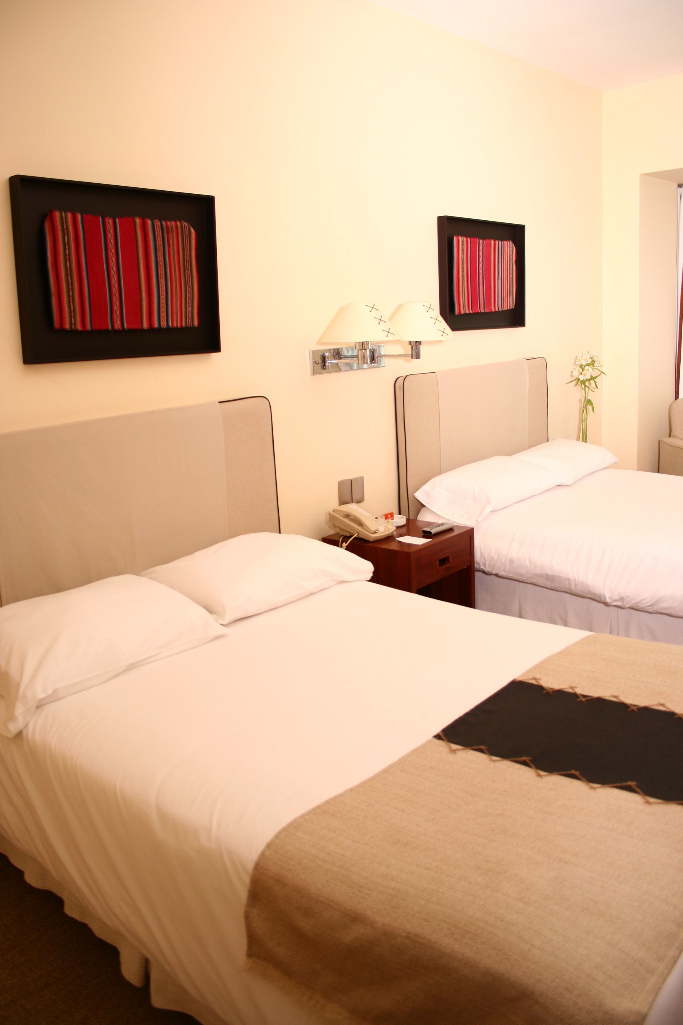 Quarto de hotel com duas camas, uma mesa de cabeceira com abajur e uma obra de arte na parede.