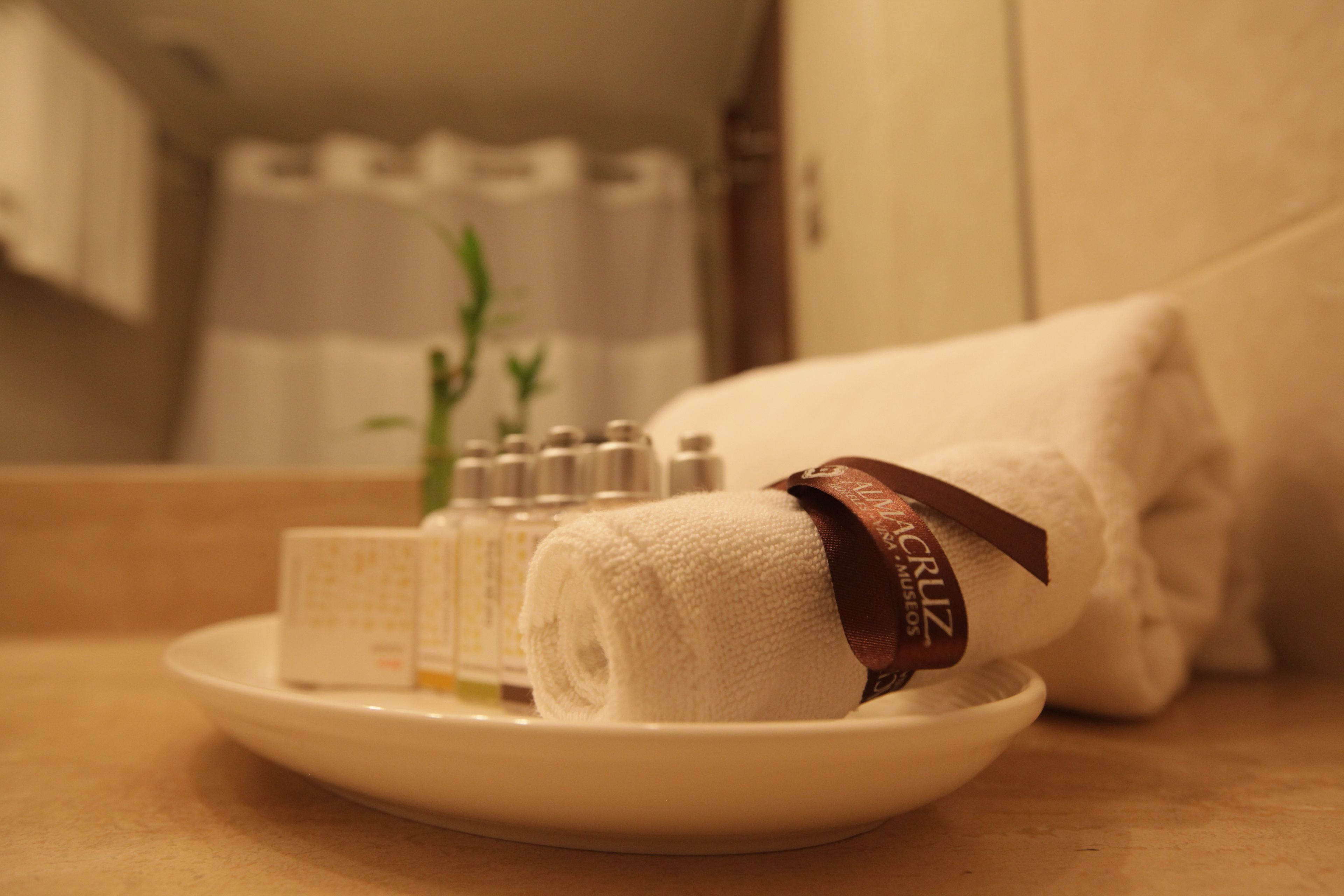 A neatly folded white towel with a brown ribbon on a white plate, a small bottle, and a glass of water on a beige countertop.