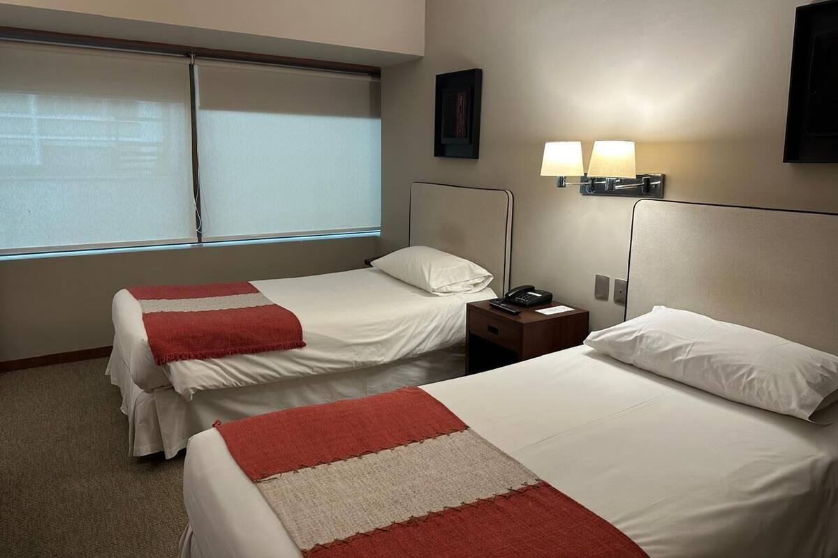 Une chambre d’hôtel avec deux lits, une table de chevet avec une lampe et une fenêtre avec des stores.