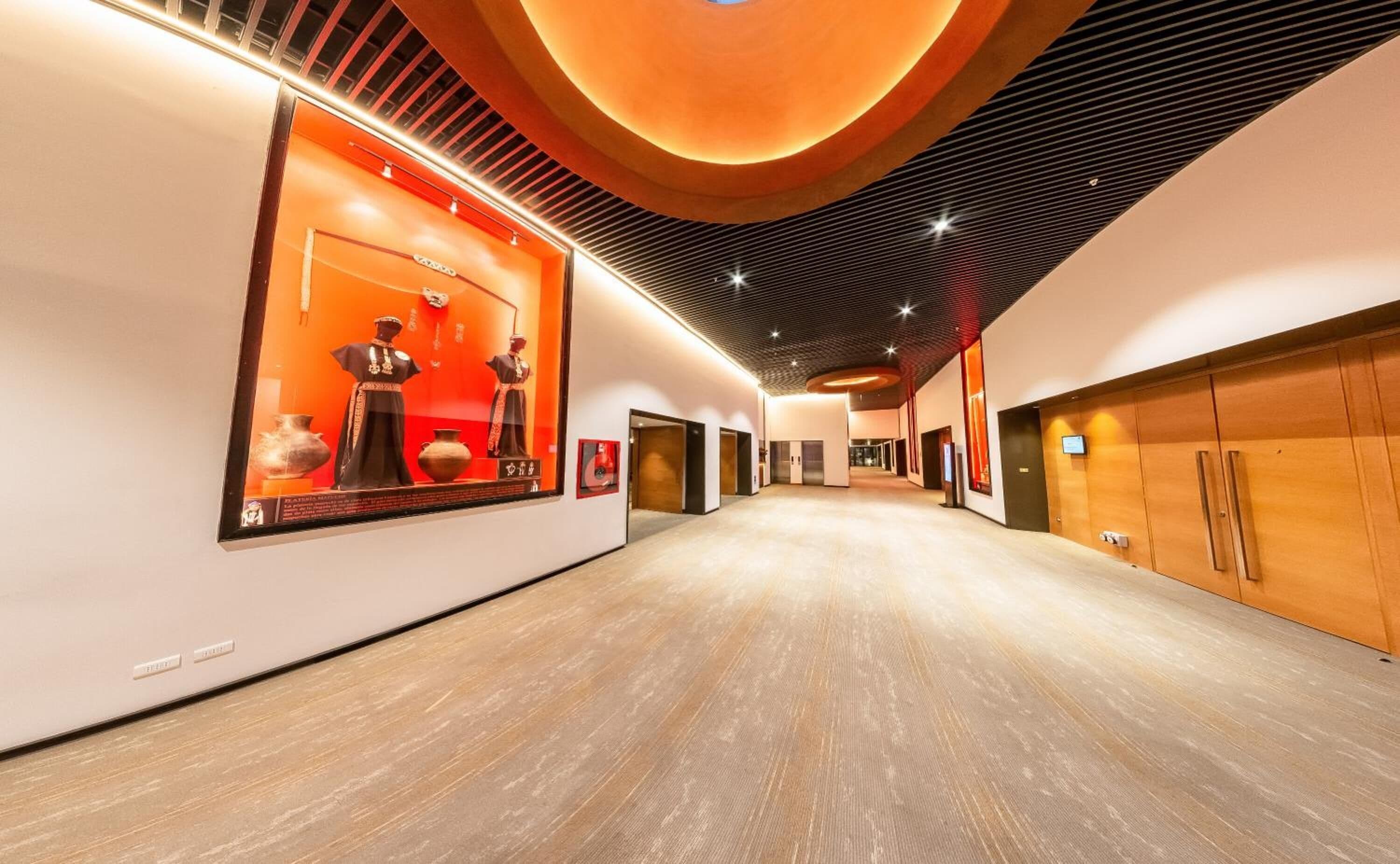 A modern hotel corridor with wooden flooring, a central display case with artifacts, and a ceiling with a circular design.