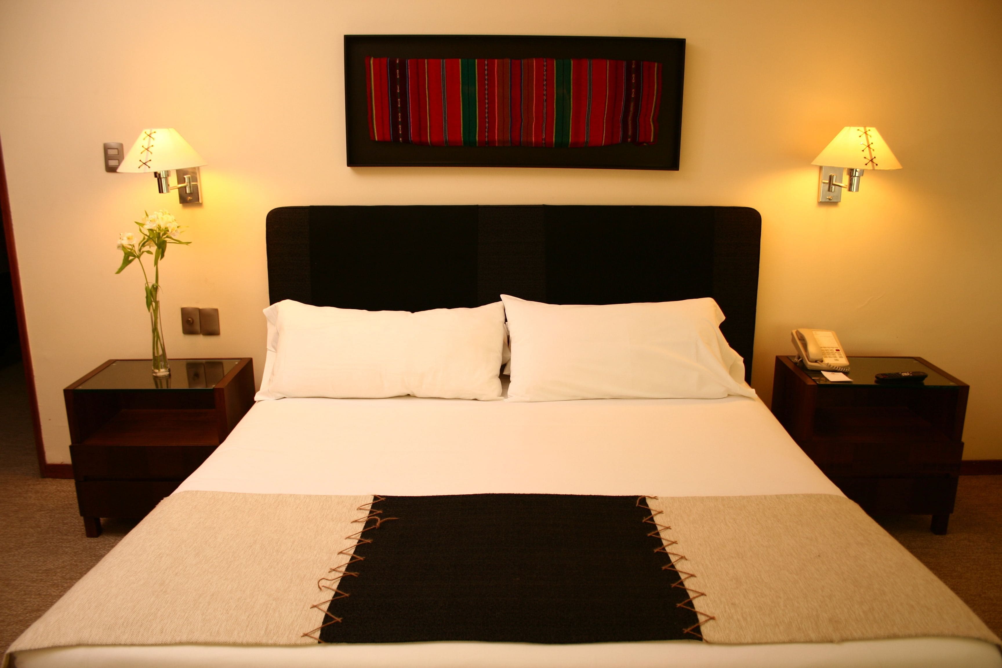 A hotel room with a large bed, two bedside tables, a nightstand, a lamp, a vase with flowers, and a framed artwork on the wall.