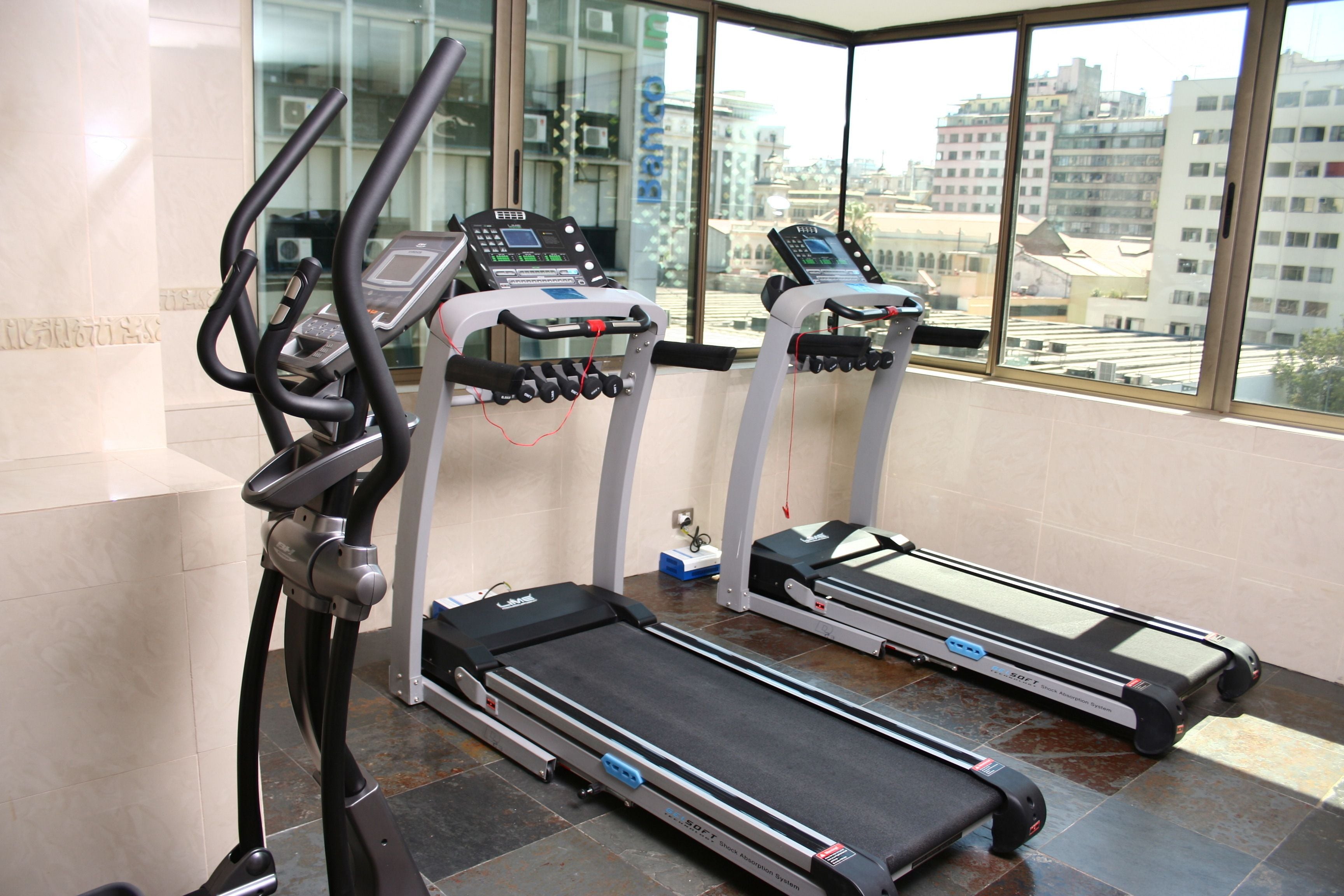 A gym with treadmills and elliptical machines, large windows with a city view, and a tiled floor.
