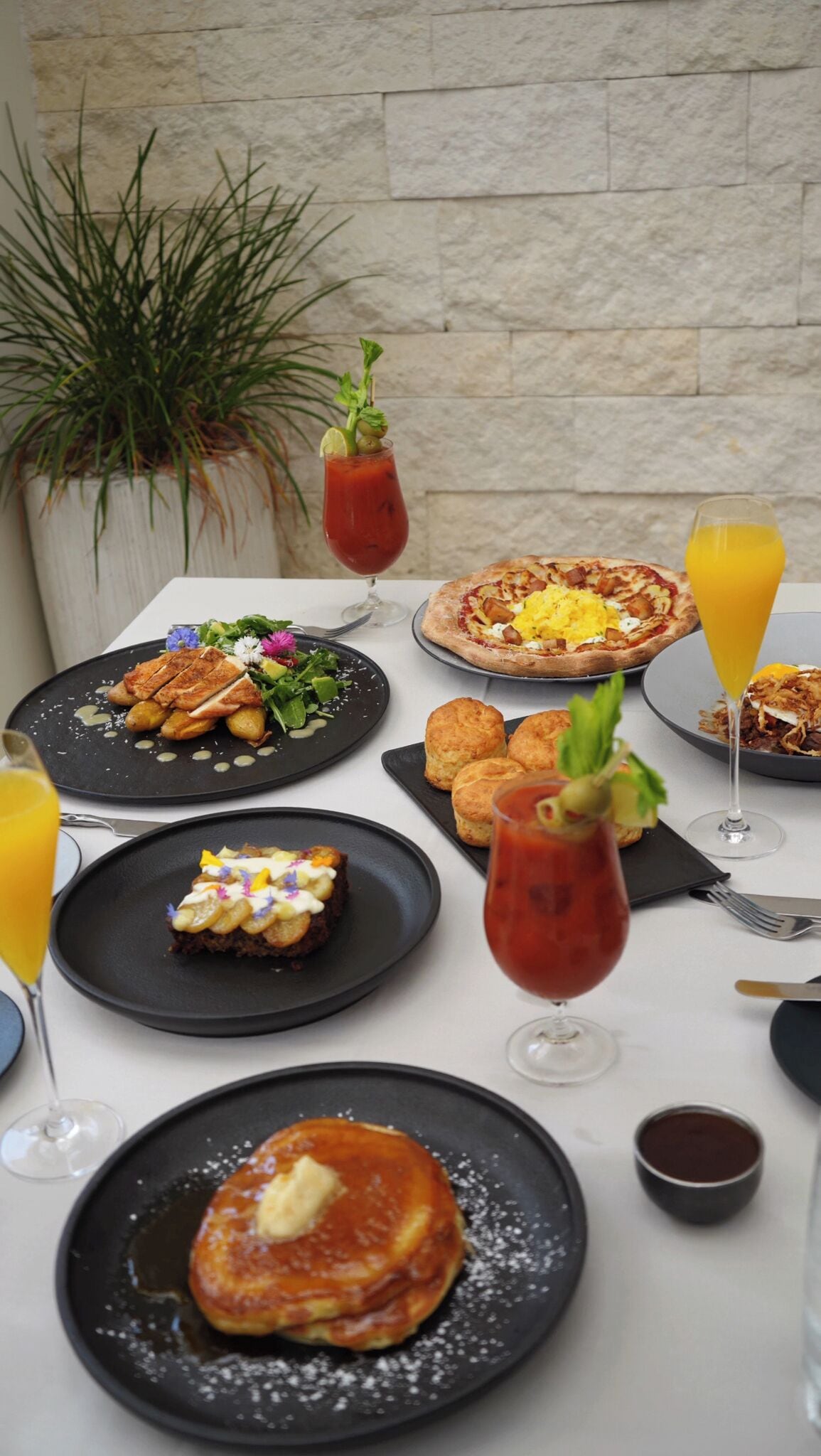 A table set with breakfast items including pancakes, a salad, and a pizza, along with drinks and a potted plant in the background.
