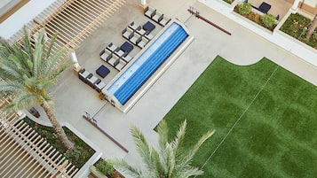 An aerial view of a hotel pool area with a long swimming pool, sun loungers, and palm trees.
