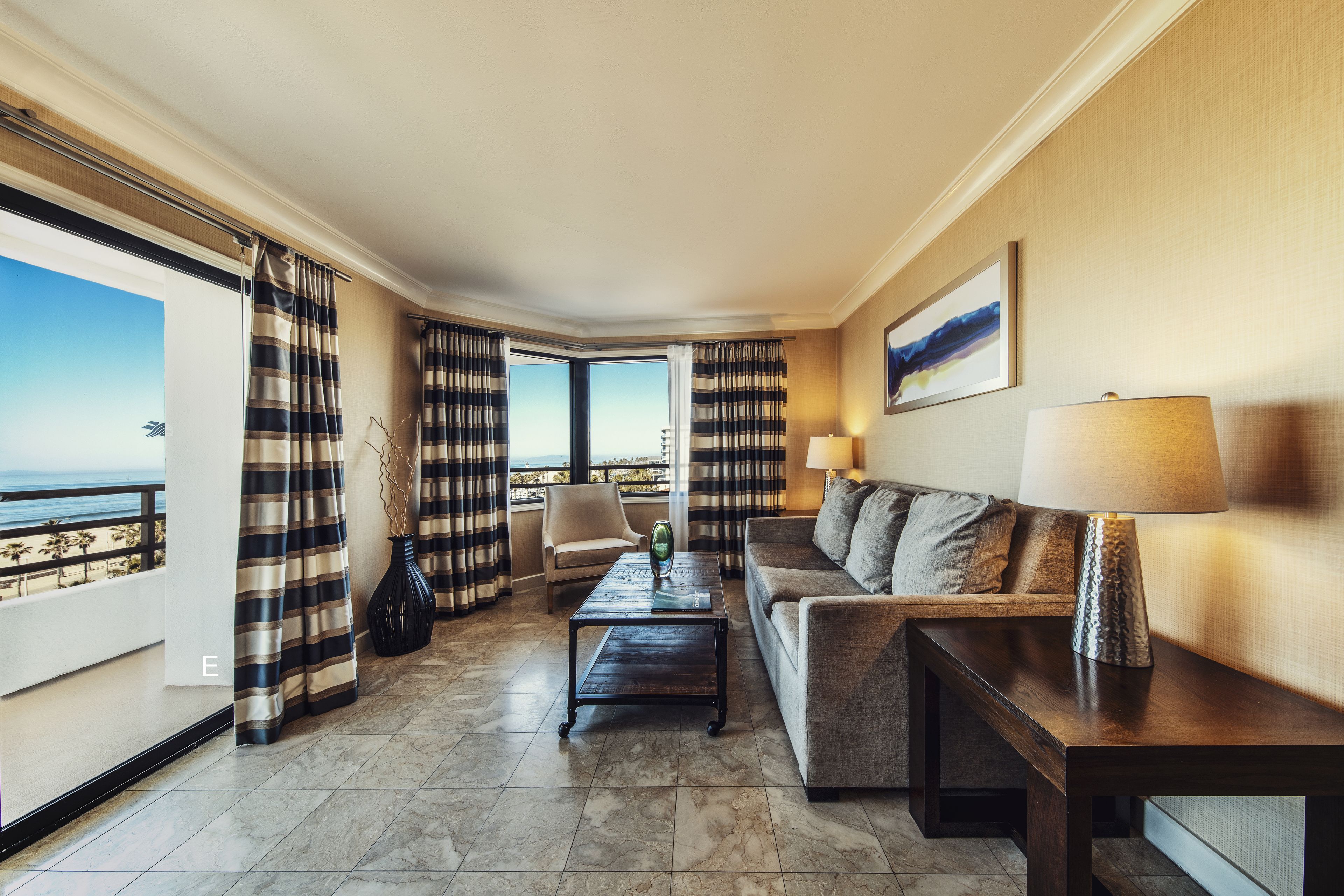 A hotel room with a sofa, a coffee table, a lamp, and a view of the ocean through large windows.
