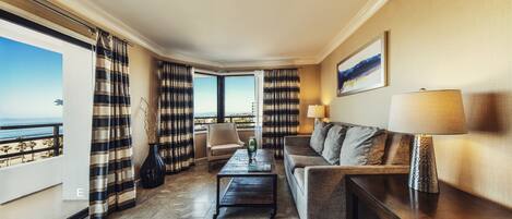 A hotel room with a sofa, a coffee table, a lamp, and a view of the ocean through large windows.