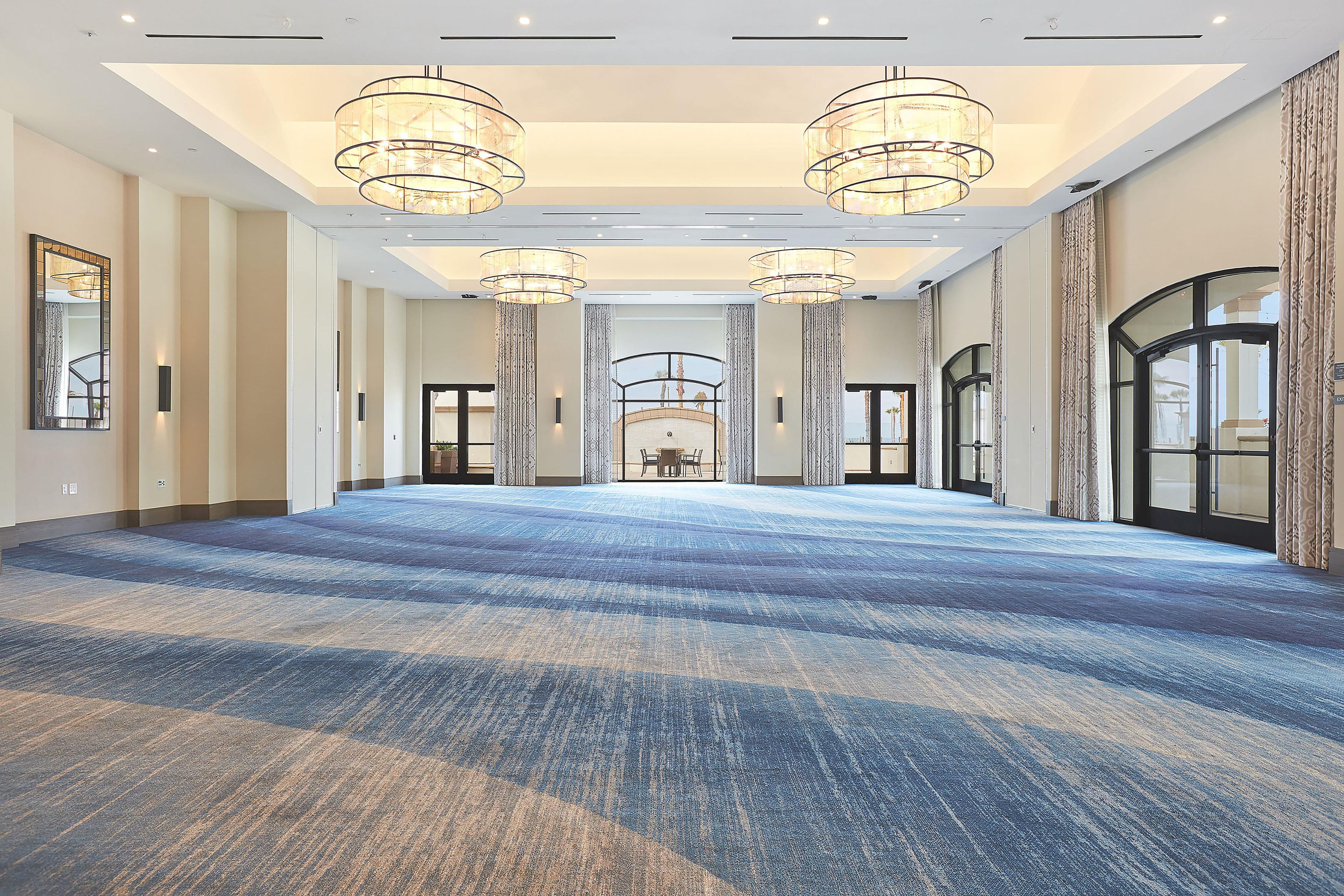 A spacious hotel lobby with a patterned carpet, large chandeliers, and a view to a dining area.