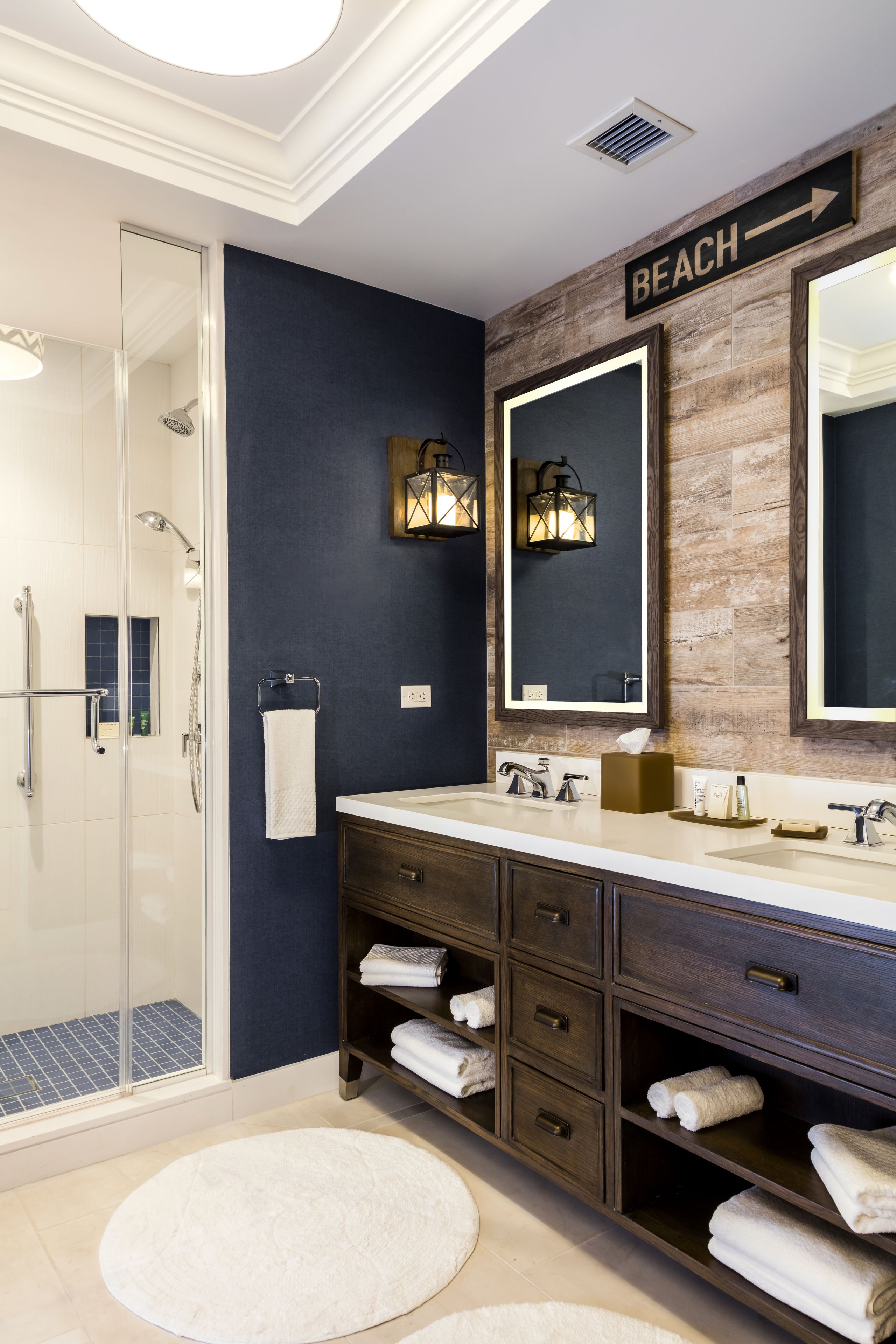 A modern bathroom with a glass shower enclosure, dark blue walls, wooden accents, and a sign indicating the direction to the beach.