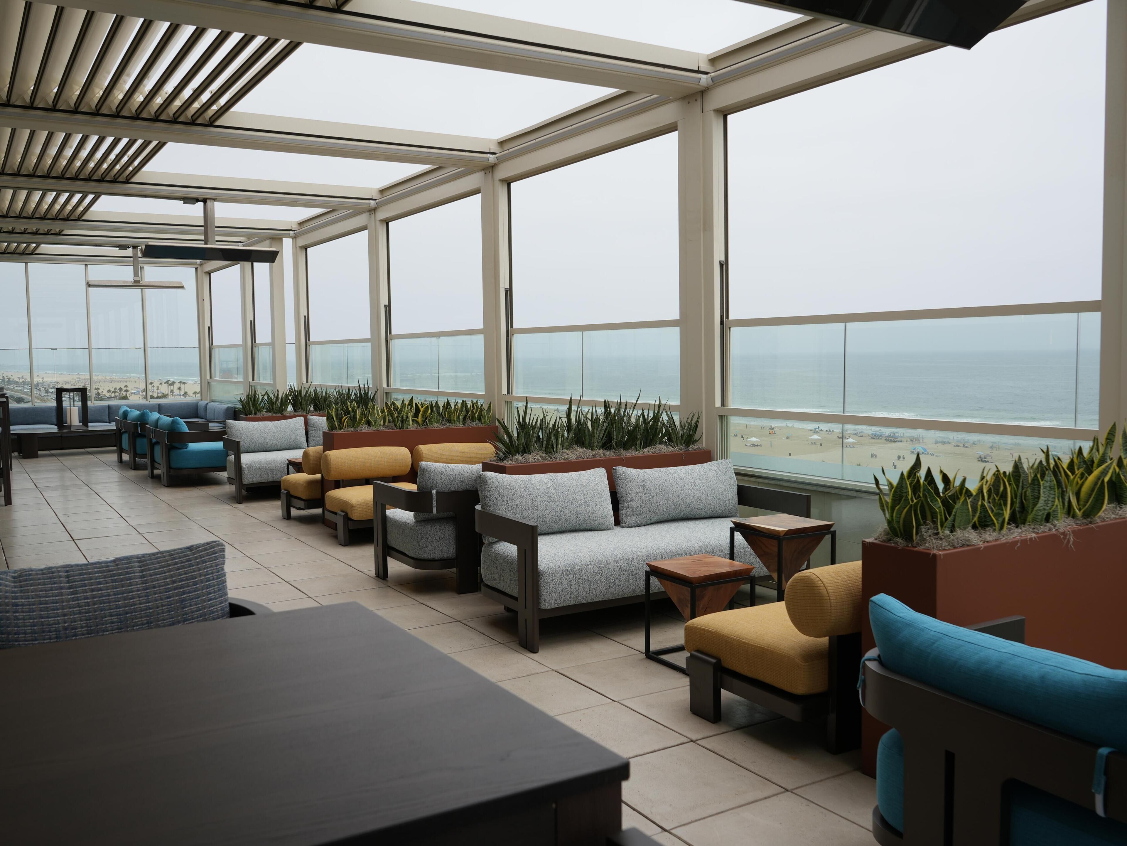 Un salon sur le toit, doté de sièges et de jardinières, de grandes fenêtres et offrant une vue sur la plage.
