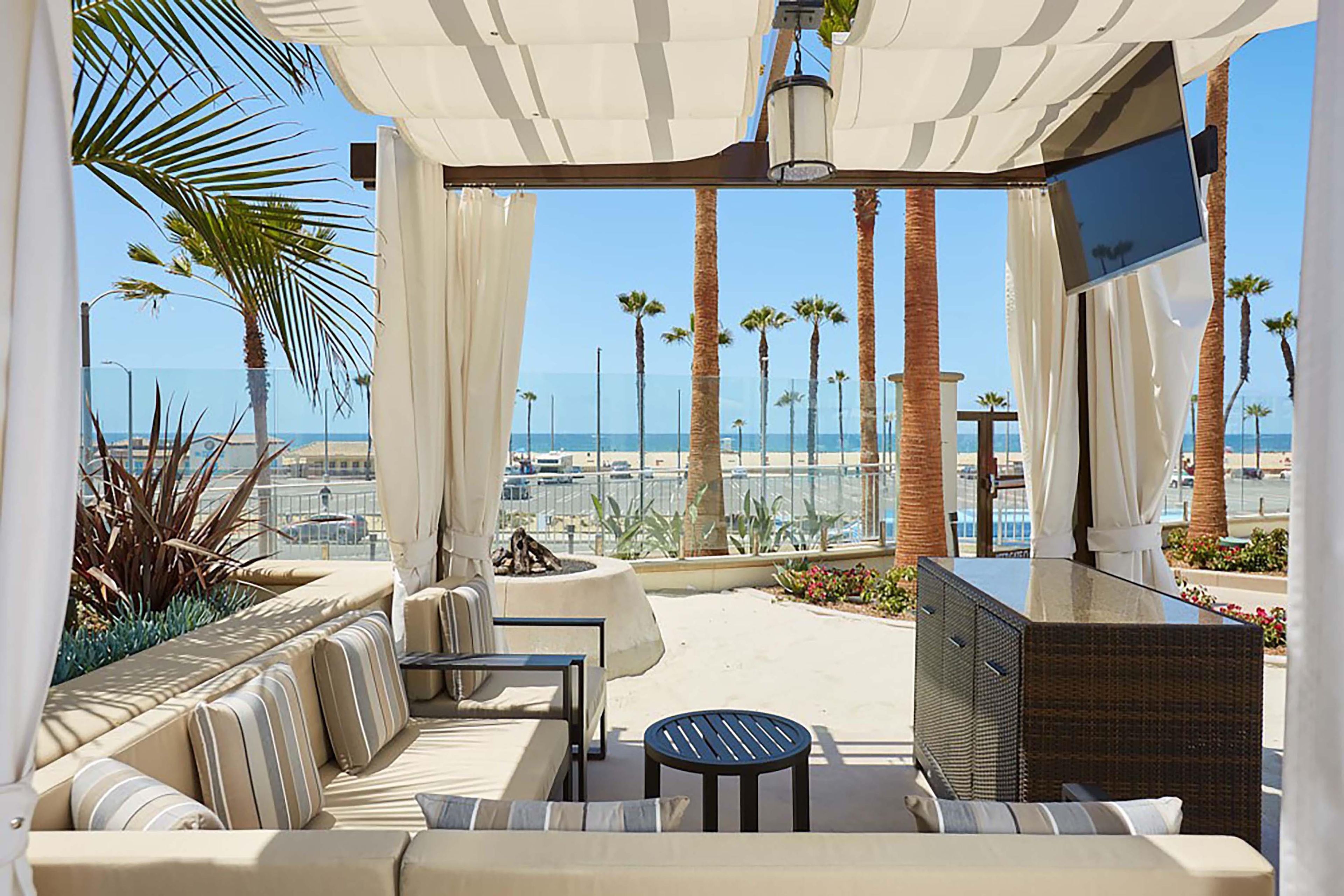 Un espace extérieur ensoleillé, aménagé avec des sièges confortables, un téléviseur et des coussins, offrant une vue sur la plage et les palmiers.