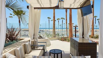 Un espace extérieur ensoleillé, aménagé avec des sièges confortables, un téléviseur et des coussins, offrant une vue sur la plage et les palmiers.