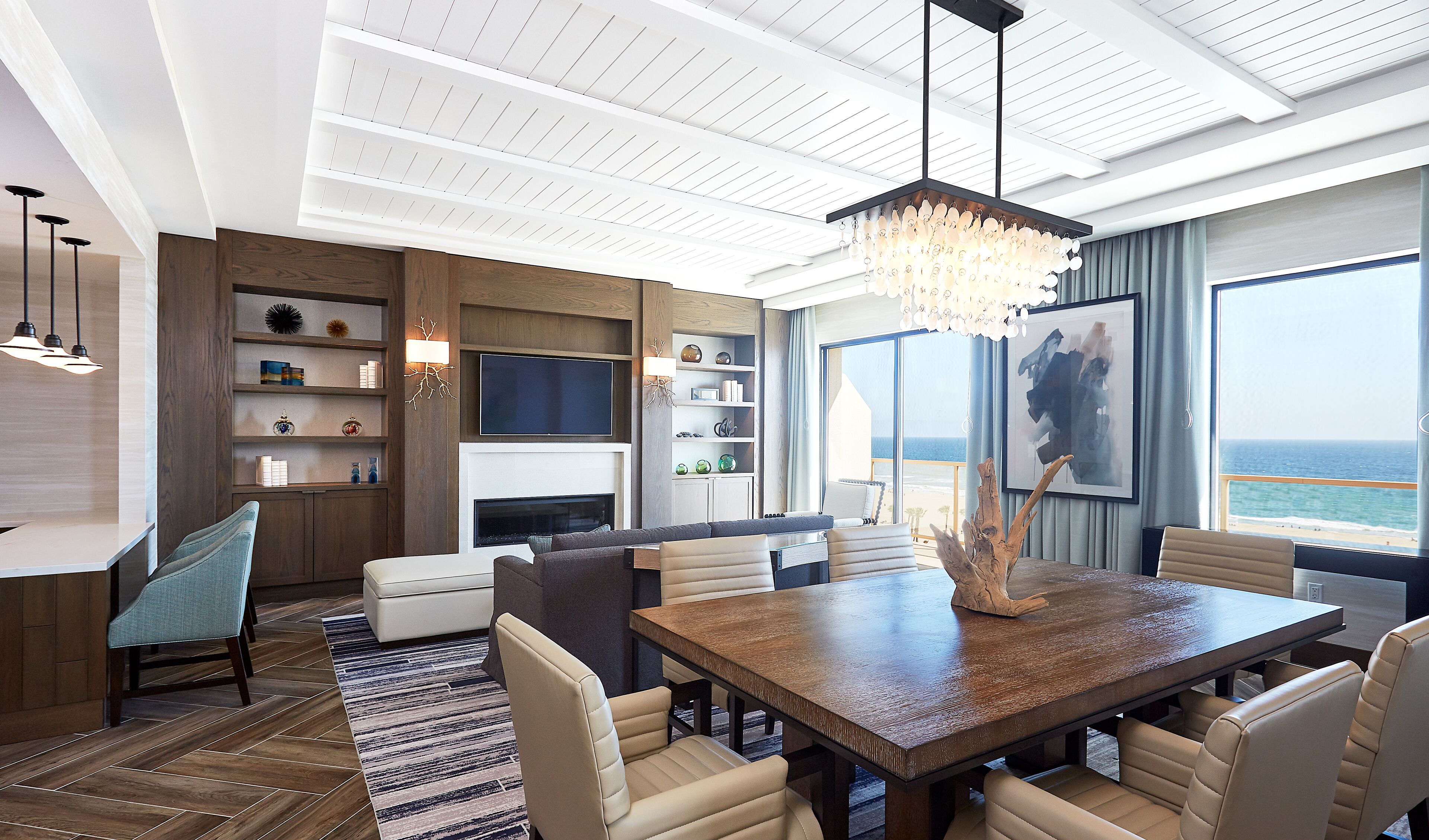 A modern hotel room with a dining area, a fireplace, a large window with a view of the ocean, and a chandelier.