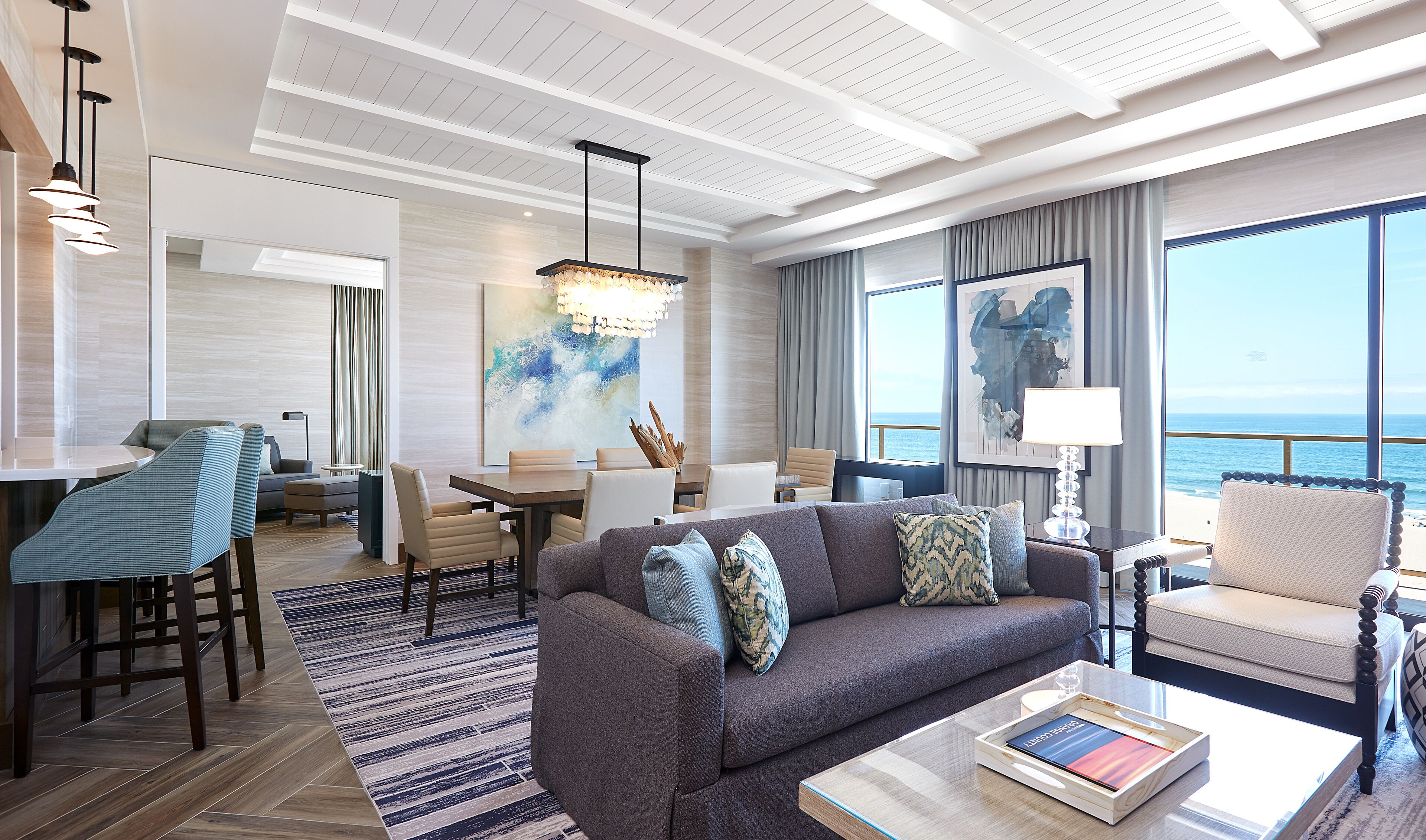 A modern living room with a sofa, coffee table, and dining area. There is a large window with a view of the ocean.