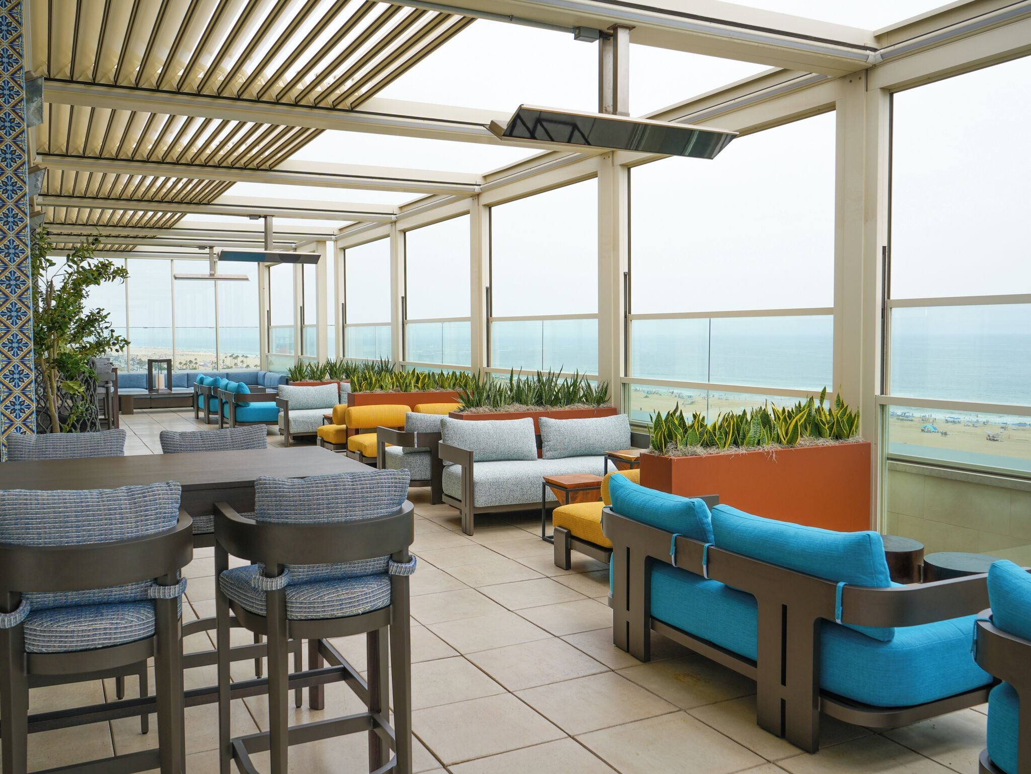 A sunlit outdoor seating area with multiple tables and chairs, large windows offering a view of the beach, and potted plants.