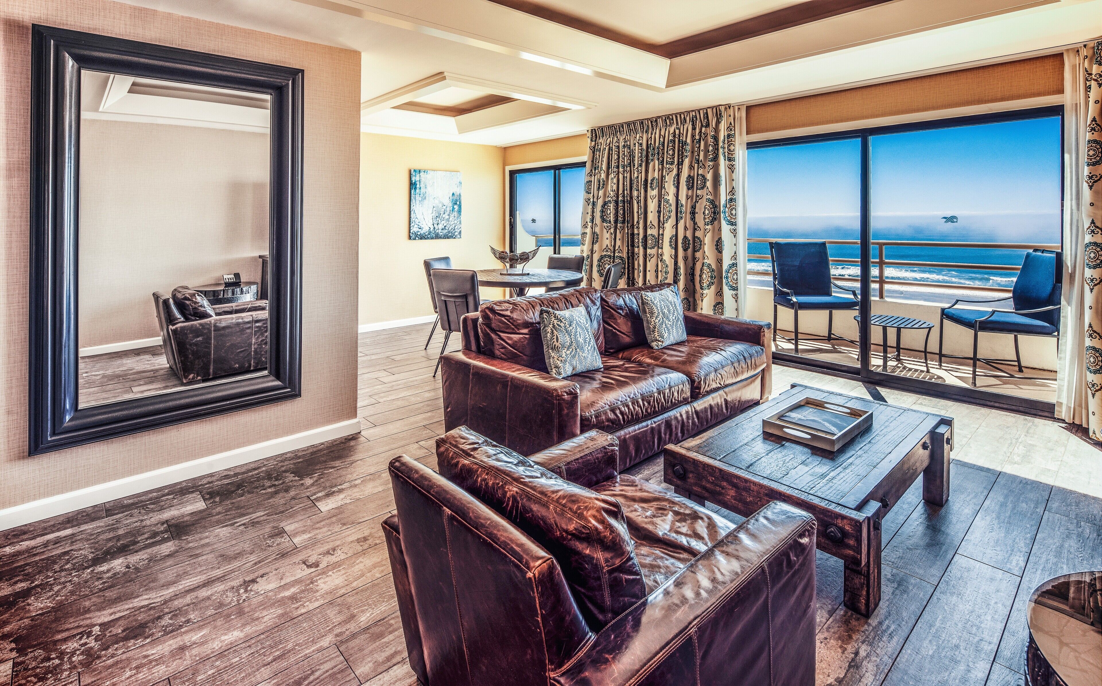 A spacious living room with a leather sofa, a coffee table, and a large mirror. There is a dining area with a table and chairs, and a balcony with a view of the ocean.