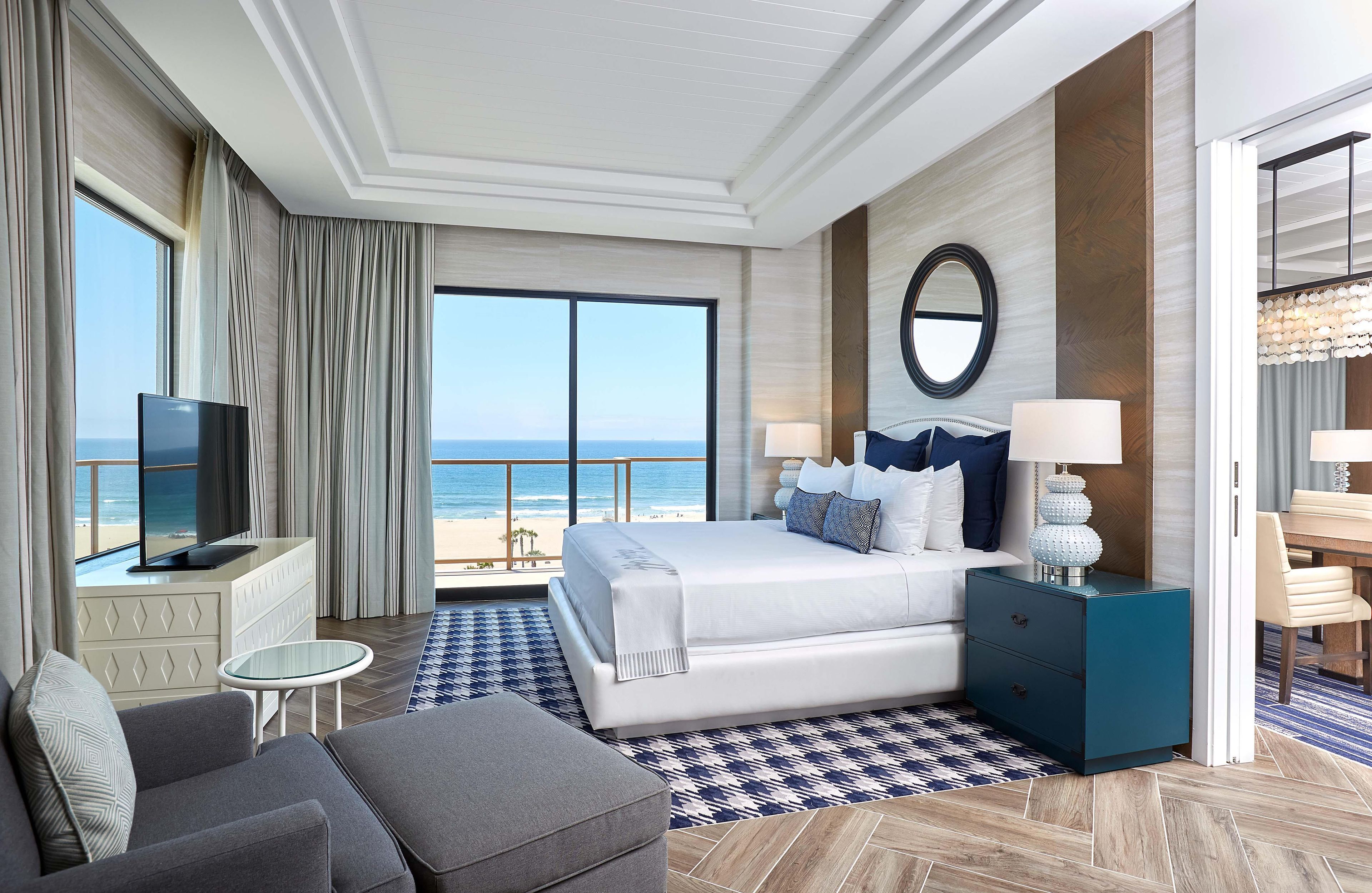 A hotel room with a large bed, a TV, a desk, and a view of the beach.
